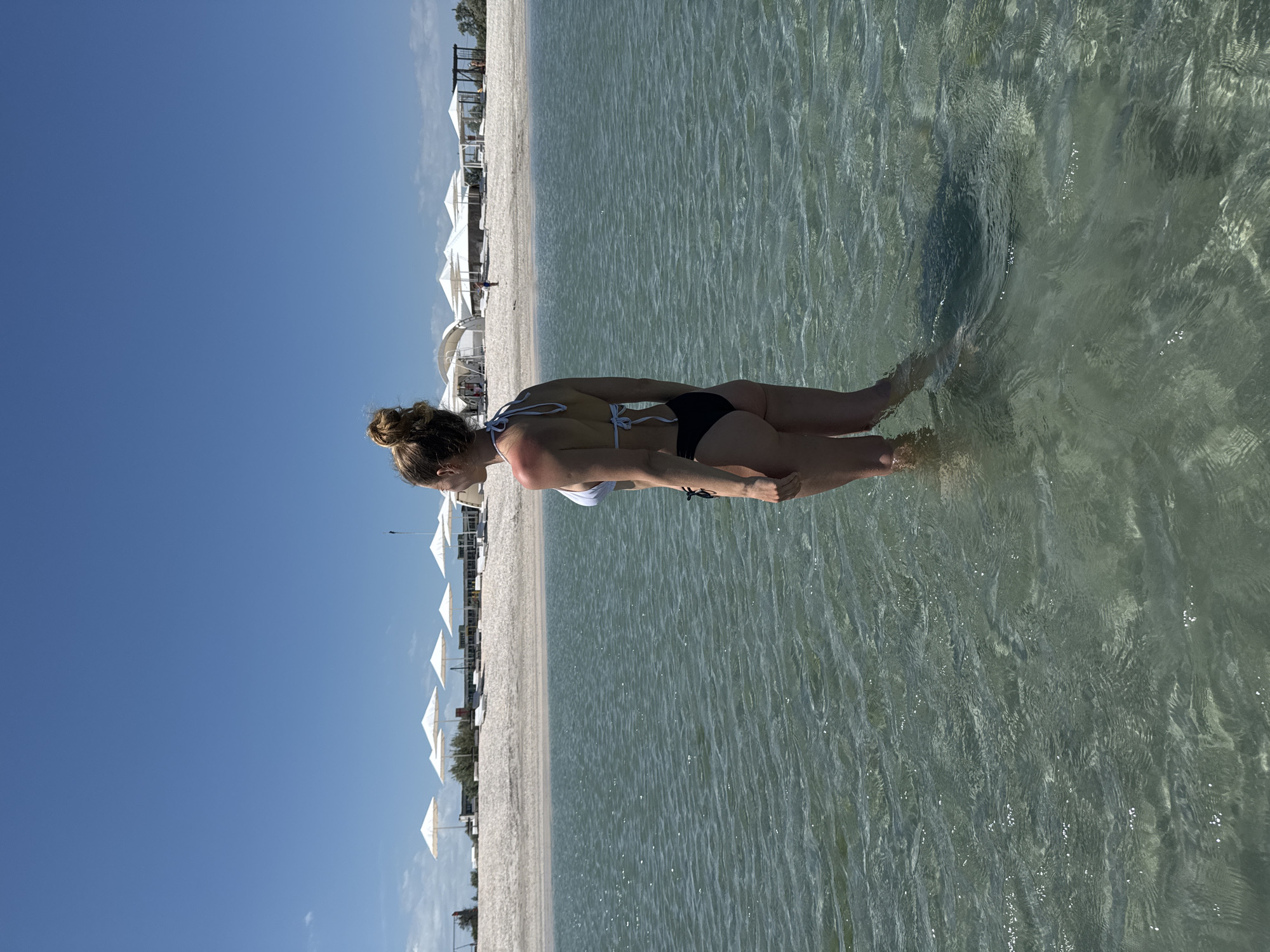 A woman is standing in the clear water of a beach in Мысовское сельское поселение, Ukraine. She is wearing a bikini. Behind her is a white sand beach with numerous white umbrellas lined up. The sky is clear and blue. The water is shallow enough to see the bottom, with small ripples on the surface.