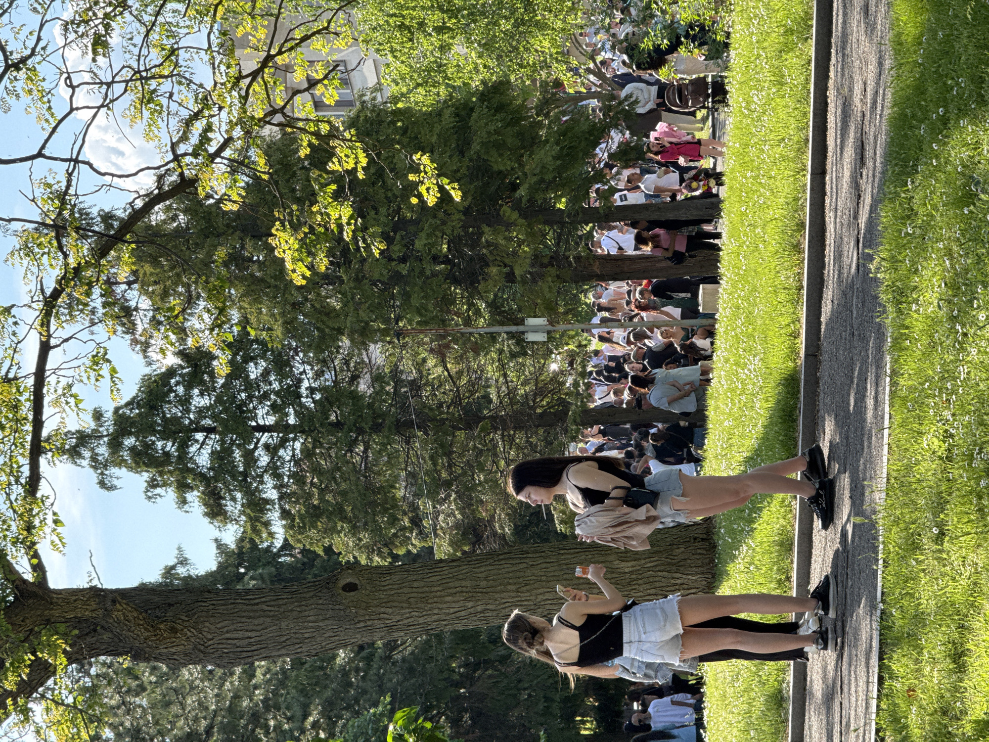 The image depicts a daytime scene in Shumen, Bulgaria. Two young women stand on a paved path that runs alongside a grassy area. Both are dressed in similar attire: black tank tops and denim shorts. One holds a drink and her phone, while the other holds a cigarette. Behind them, a large crowd of people gathers, partially obscured by trees. The background also includes buildings and greenery. The scene is bathed in sunlight.