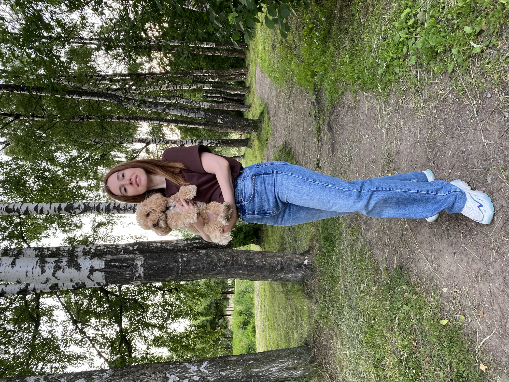 A young woman stands in a park-like setting, holding a small, light brown dog. She is wearing a brown t-shirt, blue jeans, and white sneakers. The dog is fluffy and appears to be a poodle mix. They are surrounded by birch trees with white bark, and the ground is covered in grass and a dirt path. The path curves to the right in the background, leading deeper into the wooded area. The lighting suggests it is daytime, possibly late afternoon due to the soft light filtering through the leaves. The scene is peaceful and natural, consistent with a public park or a wooded area on the outskirts of a city. Given the context, this park is likely in Kudrovo, Russia, a city known for its green spaces and parks.