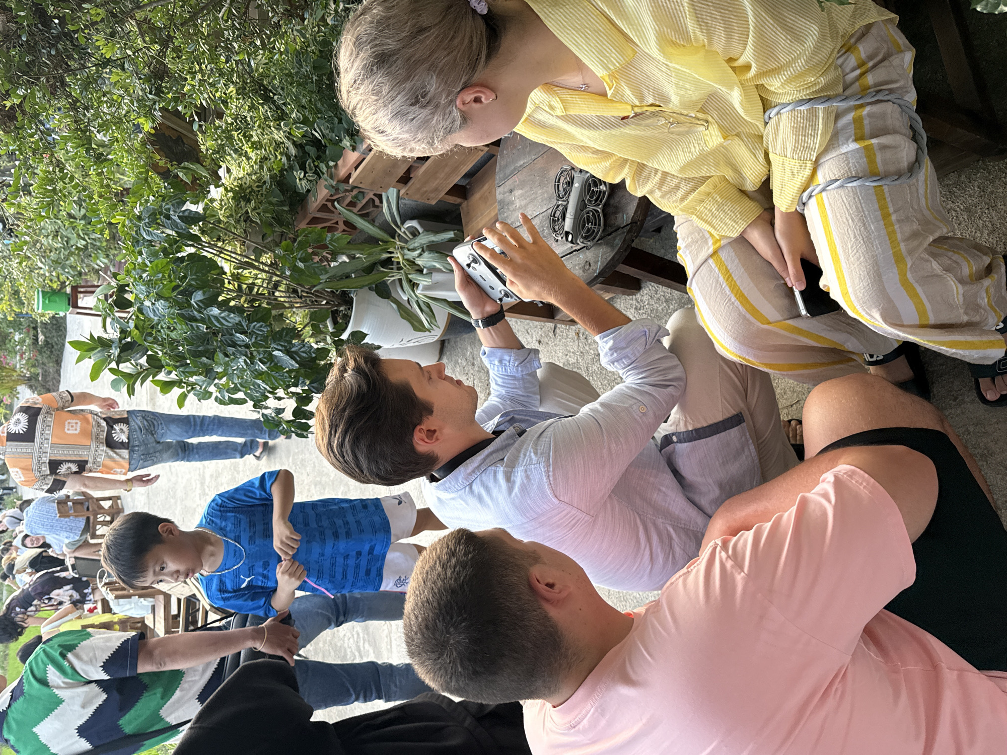 This media file captures an outdoor gathering in Diên Khánh, Vietnam, bathed in bright daylight. The scene features multiple individuals interacting in a lush, green environment.
In the central foreground, a person with dark hair, wearing a light blue striped long-sleeve shirt and light trousers, is intently focused on a white drone controller they are holding. Beside them, a person with short, light-colored hair, dressed in a yellow striped long-sleeve shirt and matching striped trousers, leans in, observing the controller with interest. A small, gray drone with four propellers is placed on a rustic wooden table next to them. The person in yellow also holds a smartphone in their lap, on the upper thigh area of the right leg, a small "FILA" logo is visible on the striped pants.
To the right, the back of a head and shoulder of a person wearing a pink t-shirt are visible. Further back, a young boy in a blue t-shirt and dark pants stands, looking towards the drone and its handlers. Several other people are partially visible in the background, including one person in an orange and black patterned shirt, suggesting a larger social event.
The setting is an outdoor space, likely a garden or patio, characterized by an abundance of green foliage, potted plants, and wooden furniture. The ground appears to be paved. The overall mood is one of engaged curiosity and social interaction, centered around the drone.