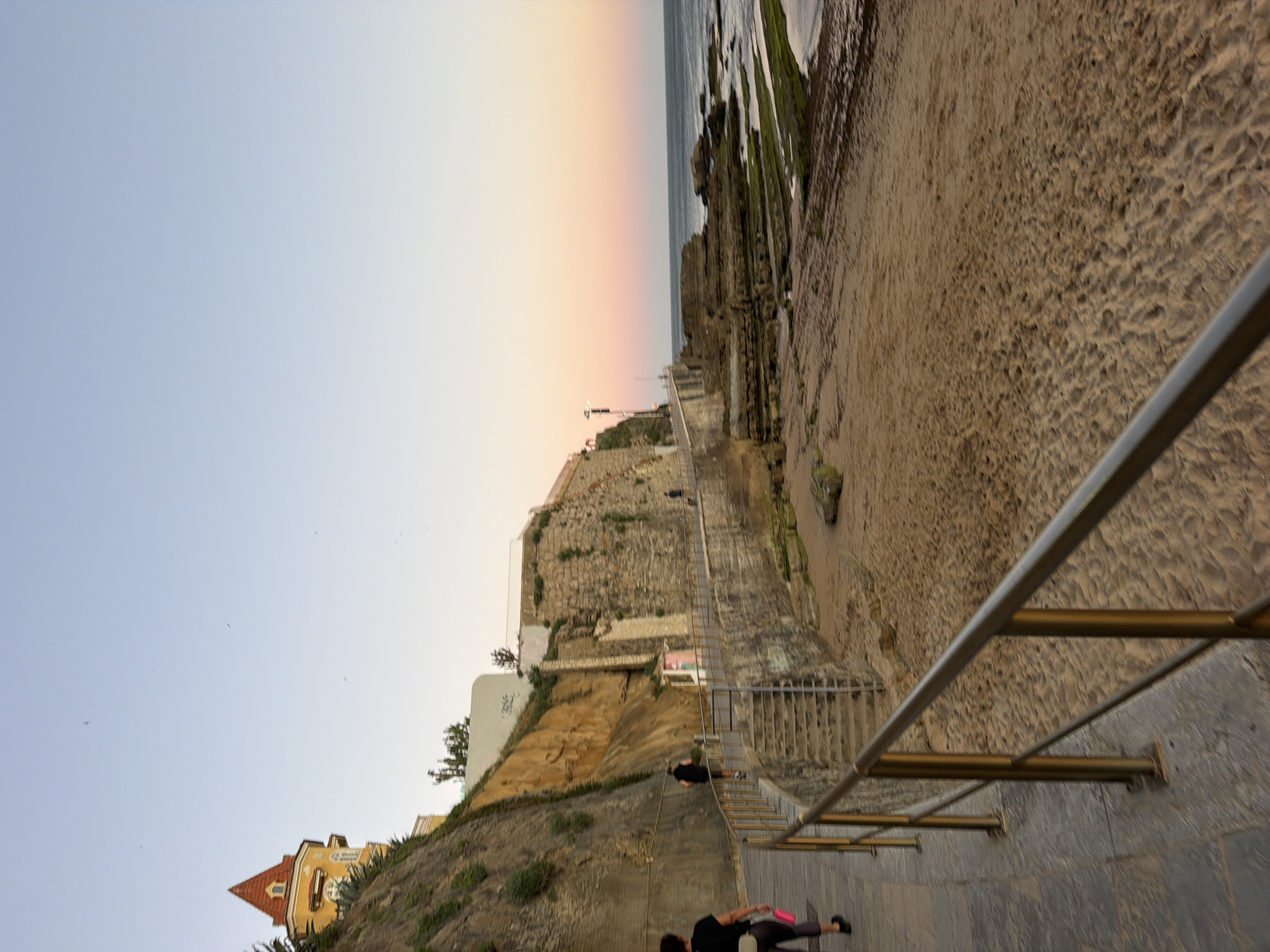 The image captures a view along a coastal path in Cascais, Portugal, during what appears to be late afternoon or early evening. A paved walkway with a metal railing runs along the left side of the frame, bordered by a steep, rocky embankment covered in sparse vegetation. Atop the embankment, a distinctive yellow building with a red, pointed roof is visible. Further along the path, stone steps lead down towards a sandy beach.
The beach itself is composed of light brown sand, with some scattered rocks and areas of seaweed. The sea is to the right, with a rocky coastline jutting out into the water. The water appears calm, and the sky above is a clear gradient of light blue to a soft pinkish hue near the horizon, indicating sunset.
There are a few individuals on the path. In the foreground, a woman with dark hair wearing dark athletic wear and carrying a pink bag walks away from the camera. Further up the path, a person in dark clothing is running. Another person is visible walking further in the distance on the path. The scene suggests a leisurely or recreational activity taking place by the coast.