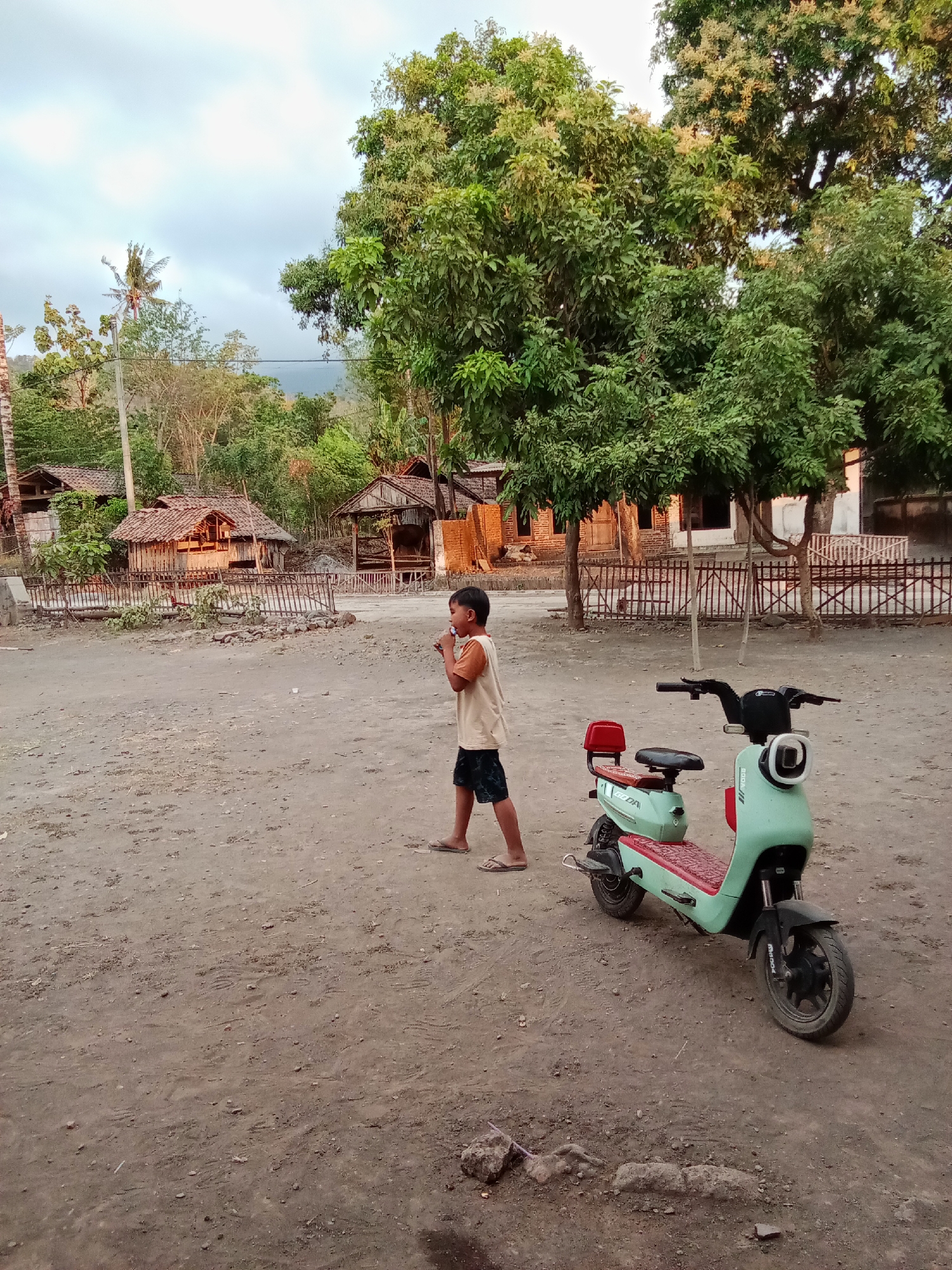 In Selowogo, Indonesia, a young boy walks across a dusty, unpaved area in front of several small, rustic buildings with thatched roofs. He is wearing a light-colored t-shirt and dark shorts. Beside him, a mint green electric scooter is parked. The background features lush green trees and a cloudy sky.
