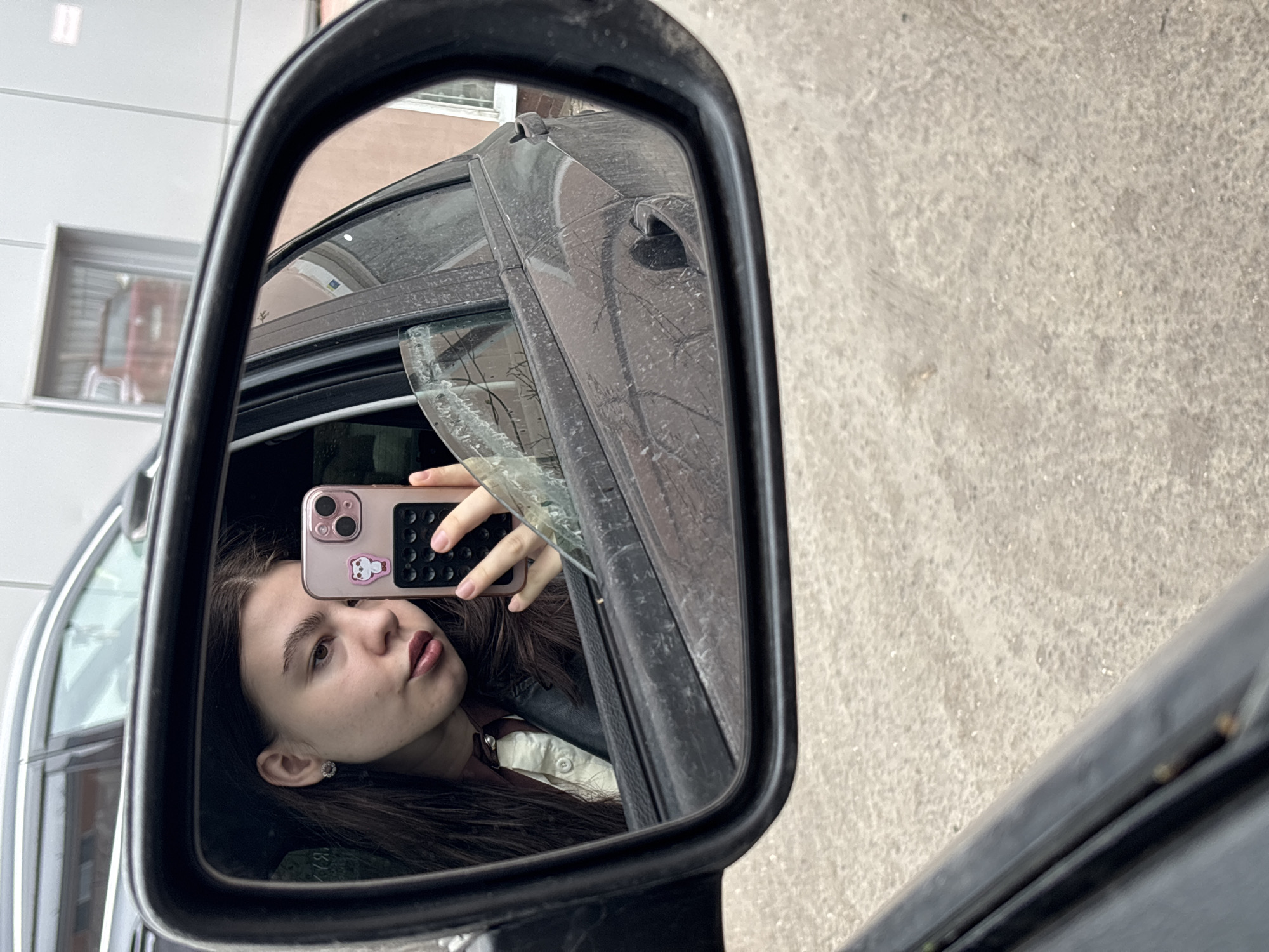 The image shows a reflection in a car's side mirror. A young woman is taking a selfie using a phone with a pink case and a small bear sticker. The car's door and window frame are visible. In the background, a building with a window is reflected. Based on the architectural style of the building, the location is likely Perm, Russia. The exterior of the car and the side mirror are dirty.