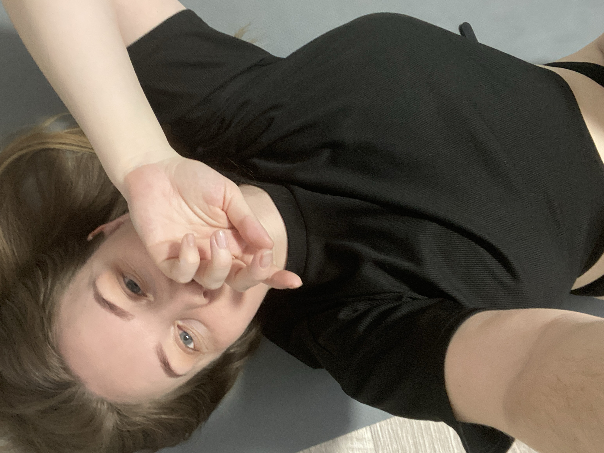 An indoor scene, presumably in Noviy, Russia, depicts an adult female positioned supine on a grey exercise mat. The individual is captured in a selfie-style photograph, looking directly into the camera. She presents with pale skin, blue eyes, and light brown hair. Her attire consists of a black short-sleeved, ribbed top worn over a black undergarment. The subject's left hand, featuring light-colored manicured nails, is partially raised, obscuring her forehead and the upper portion of her eyes. The immediate environment includes a plain grey wall in the background and a light-colored floor, consistent with wood or laminate, visible to the right of the mat.