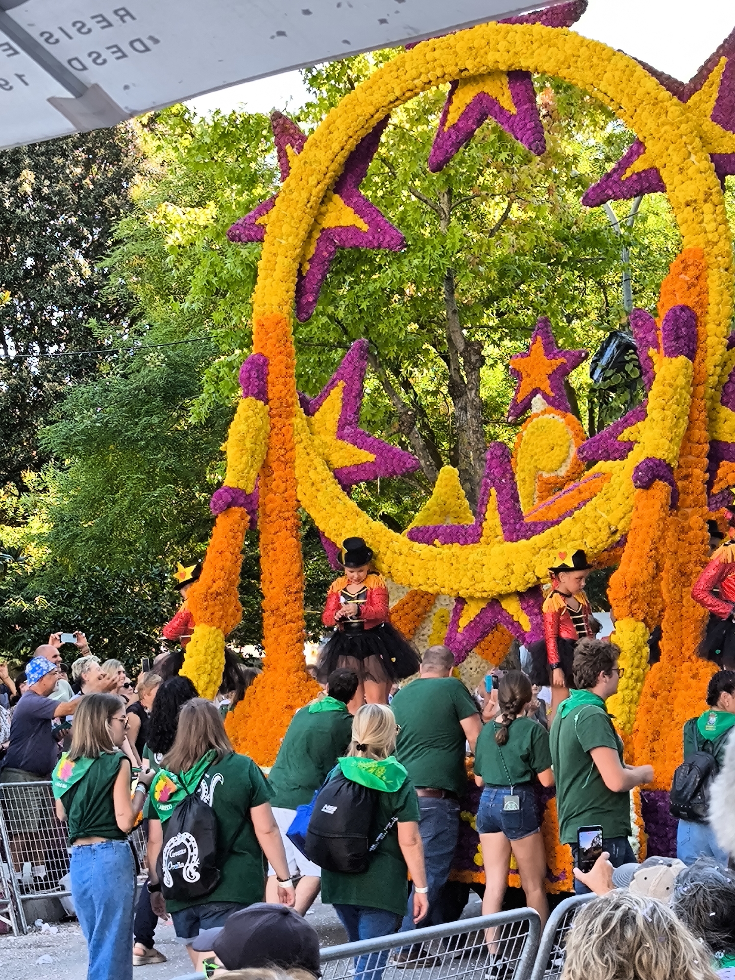 The image depicts a vibrant floral float at an outdoor event in Laredo, Spain. The float is a large, circular structure adorned with yellow, orange, and purple flowers, forming star shapes and circular patterns. Several young people in costumes, possibly performers, are visible on the float. Some are wearing red and black jackets with gold trim and black tutus, while others have on black hats. In the foreground, a crowd of spectators is gathered, many holding up cameras to take pictures. They are dressed in casual clothing, and some are wearing green shirts with white logos. The background consists of lush green trees, suggesting a sunny day. A white banner with black text is visible at the top of the image, and a metal fence separates the crowd from the float.