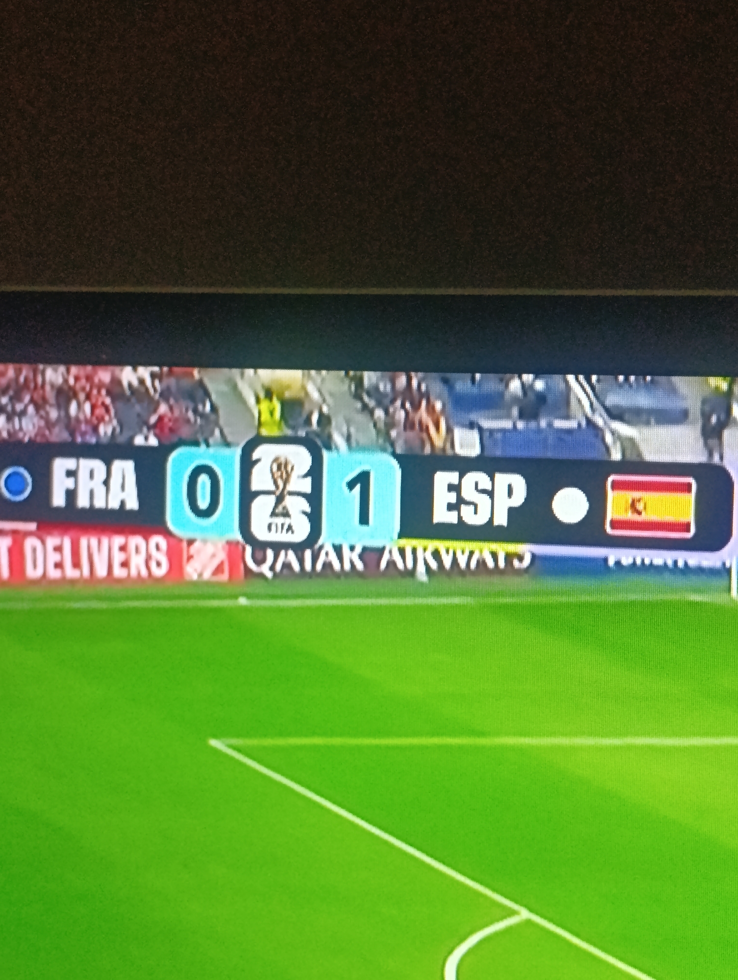 The image shows a soccer match scoreboard displaying "FRA 0 - 1 ESP". The scoreboard is positioned on a large screen, likely at a stadium. The screen shows the flags and abbreviations for France and Spain, with "FRA" representing France and "ESP" representing Spain. The score indicates France has 0 goals and Spain has 1 goal. Below the score, there is advertising text that reads "QATAR AIRWAYS". The background shows a blurred image of a crowd in a stadium and a green soccer field. The image quality is poor, with visible pixelation and noise, suggesting it was taken with a camera or phone at a distance. There is no visible indication of Osh City, Kyrgyzstan in the image.