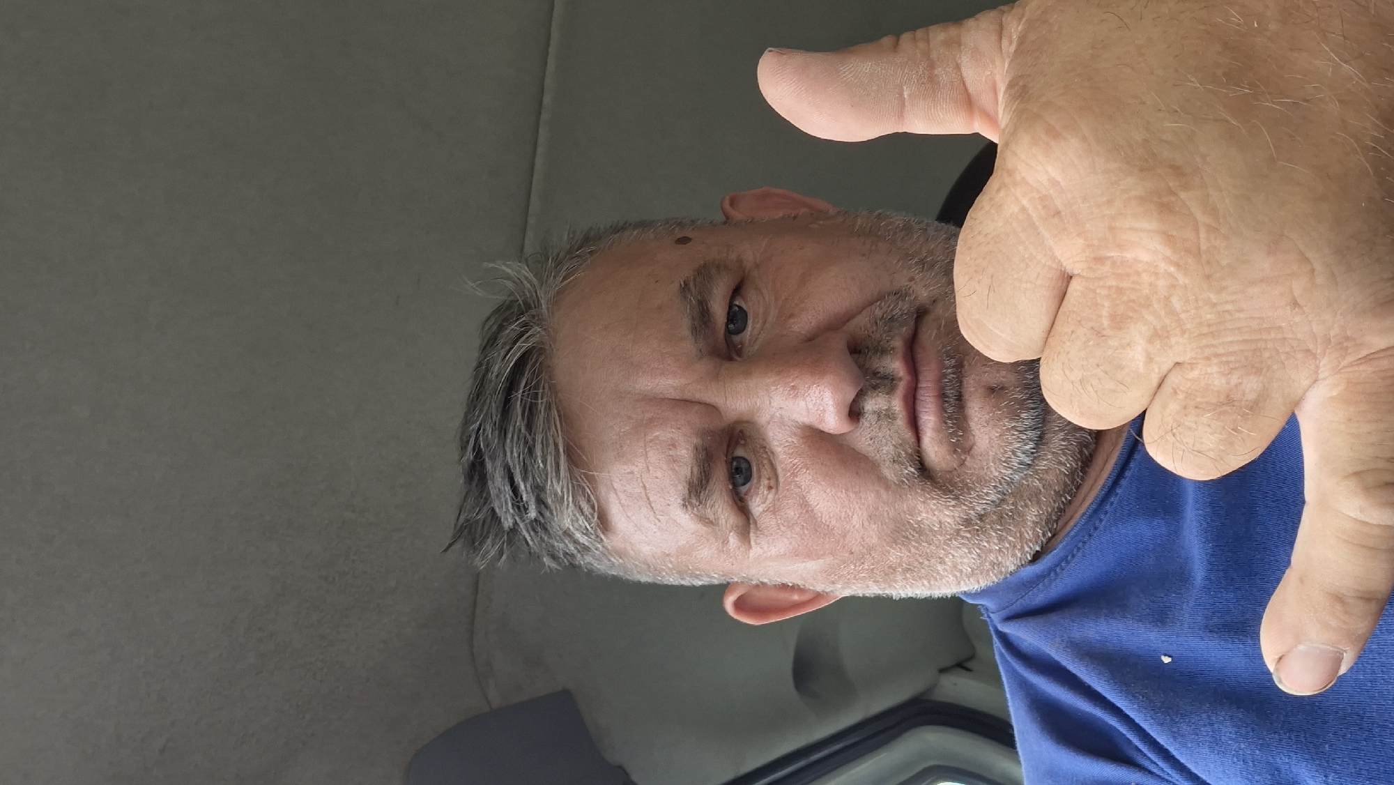 A man with graying hair and a beard is sitting in a vehicle, likely the interior of a car or van. He is wearing a blue t-shirt and giving a thumbs-up gesture with his right hand. His left hand is partially visible in the lower left corner, forming a "shaka" or "hang loose" sign. The man's facial expression is neutral, and his eyes are looking directly at the camera. The background appears to be the interior of a vehicle, with gray upholstery visible. The lighting suggests an overcast day. There are no clear indicators of the specific location within Hîncești, Moldova.