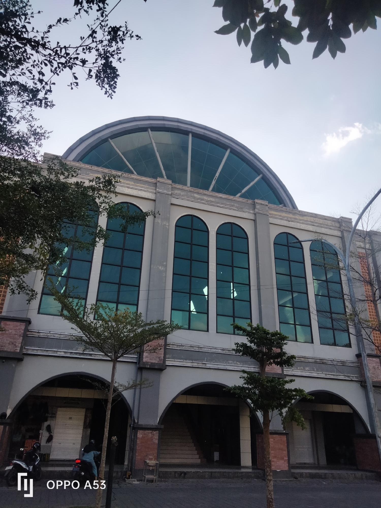 This is a medium-shot, eye-level, exterior photograph of a multi-story building with a large, arched glass roof. The building has a light gray facade with large, arched windows featuring dark frames. Several trees are visible in front of the building, some with dark green foliage and others with sparser branches. The ground in front of the building is paved. In the foreground, two motorcycles are parked, and two individuals are on one of the motorcycles, appearing to be riding away from the camera. The sky above is partly cloudy and light blue. The image also contains a watermark in the lower-left corner indicating "OPPO A53". The context provided suggests this location is in the city of Trenggalek, Indonesia.