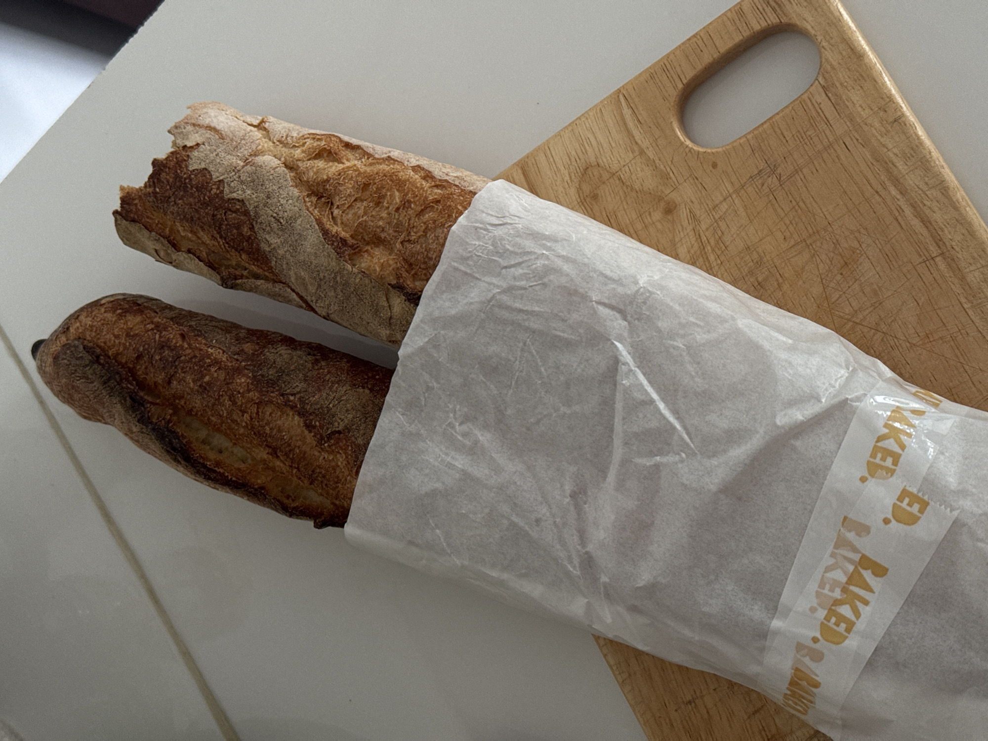Two baguettes are wrapped in white paper, with a strip of tape reading "BAKED" repeatedly across it. The baguettes are placed on a wooden cutting board. The scene is indoors, on a white countertop, likely in a kitchen. The image does not contain enough information to determine the location in the city of North Kuta, Indonesia.