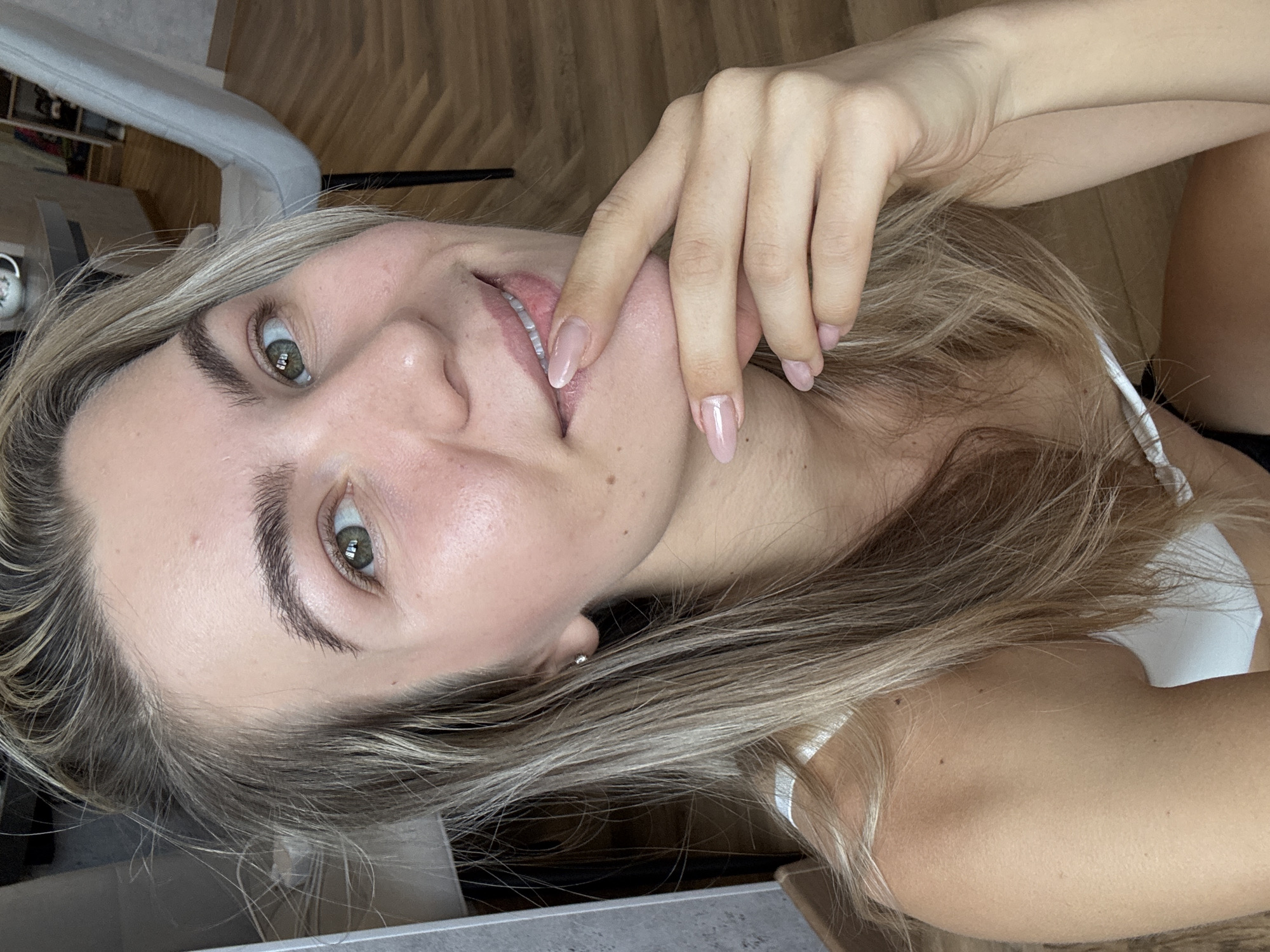 A close-up, slightly blurred image depicts a fair-skinned woman with light blonde hair. She is oriented on her side, looking directly at the camera. Her left hand is raised, with the fingertips near her mouth. She has green eyes, and her eyebrows are well-defined. Her nails appear to be manicured with a pale pink polish. She is wearing a white garment. The background features a wooden floor and a grey piece of furniture. The location is Unknown, Unknown.