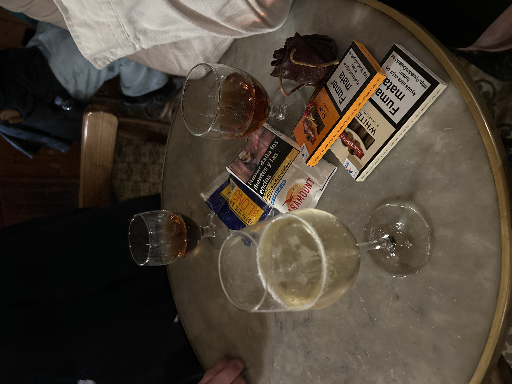 The image shows a top-down view of a round marble table with a gold trim, cluttered with various items. There are three glasses of different sizes and shapes, two containing amber-colored liquids, presumably alcoholic beverages, and one containing a lighter, bubbly drink.
Several packets of cigarettes or cigars are scattered around the glasses, including brands like "Fumar mata" and "Paramount". A small brown leather pouch with drawstrings is also visible.
Clothing and furniture are partially visible around the edge of the table, suggesting people are seated around it. The presence of cigarettes, cigars, and alcoholic beverages indicates a social gathering or a moment of relaxation.
Without more contextual details, it's difficult to pinpoint the exact location in Barcelona. The presence of marble tables and the style of the items might suggest a bar, café, or restaurant, common in Barcelona.