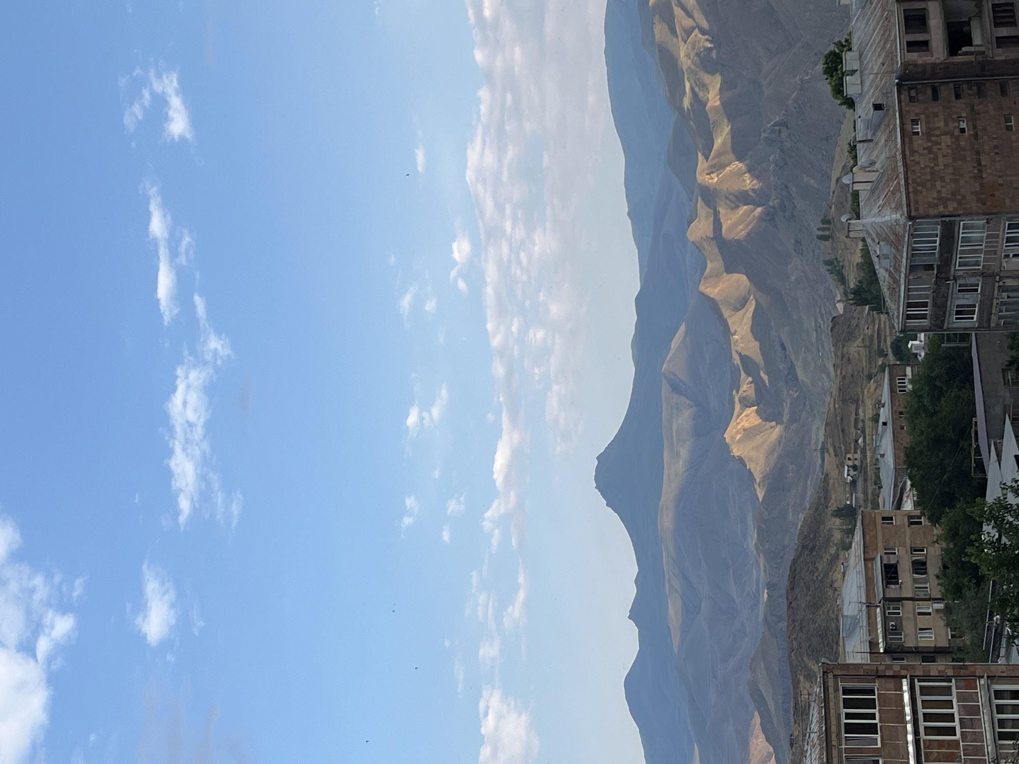 The image depicts a panoramic view of the city of Gladzor, Armenia, with a prominent mountain range in the background. The foreground is occupied by residential buildings, characterized by their concrete structures and multiple stories. The sky is a vibrant blue, adorned with scattered white clouds. The mountains, bathed in the soft light of what appears to be either early morning or late afternoon, showcase a rugged terrain with varied textures and shadows. Patches of vegetation are visible on the slopes of the hills closer to the city. A few birds are captured in flight against the expansive sky. The overall scene suggests a peaceful, somewhat remote urban environment set against a dramatic natural landscape.