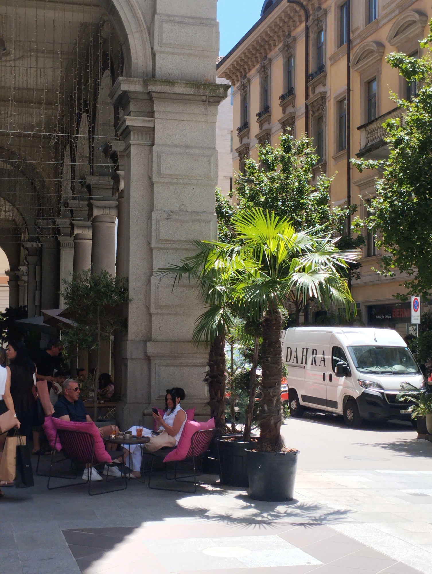 In the city of Lugano, Switzerland, on a sunny day, a street scene unfolds under the arches of a building. Two people are seated at a small round table, facing each other. The person on the left, a man wearing sunglasses and a dark blue t-shirt, is looking towards the right. The person on the right, a woman with dark hair, is wearing a white top and looking down at her phone. Both are seated in pink chairs with cushions. A white van with "DAHRA" written on its side is parked on the street in the background, partially obscuring a storefront. Lush green trees, including palm trees, are visible throughout the scene, casting shadows on the paved ground. The architecture of the building features stone columns and decorative elements.