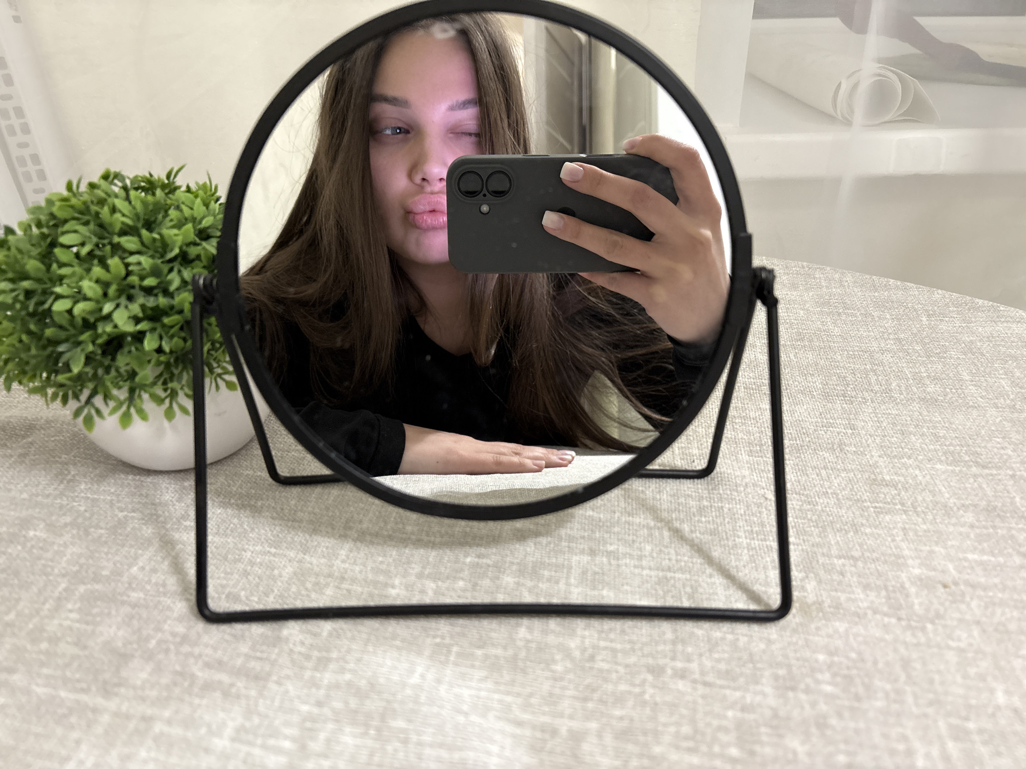 A young woman is seen in the reflection of a circular mirror with a black metal frame, positioned on a table covered with a neutral-toned, textured tablecloth. She is holding a smartphone, possibly an iPhone, and taking a selfie with a wink and pursed lips. Her hair is long and brown. A small potted plant is placed next to the mirror on the table. The background suggests an interior space with a roll of white material visible on a shelf. The location of the photo is identified as Chaikovsky, Russia, based on available information.