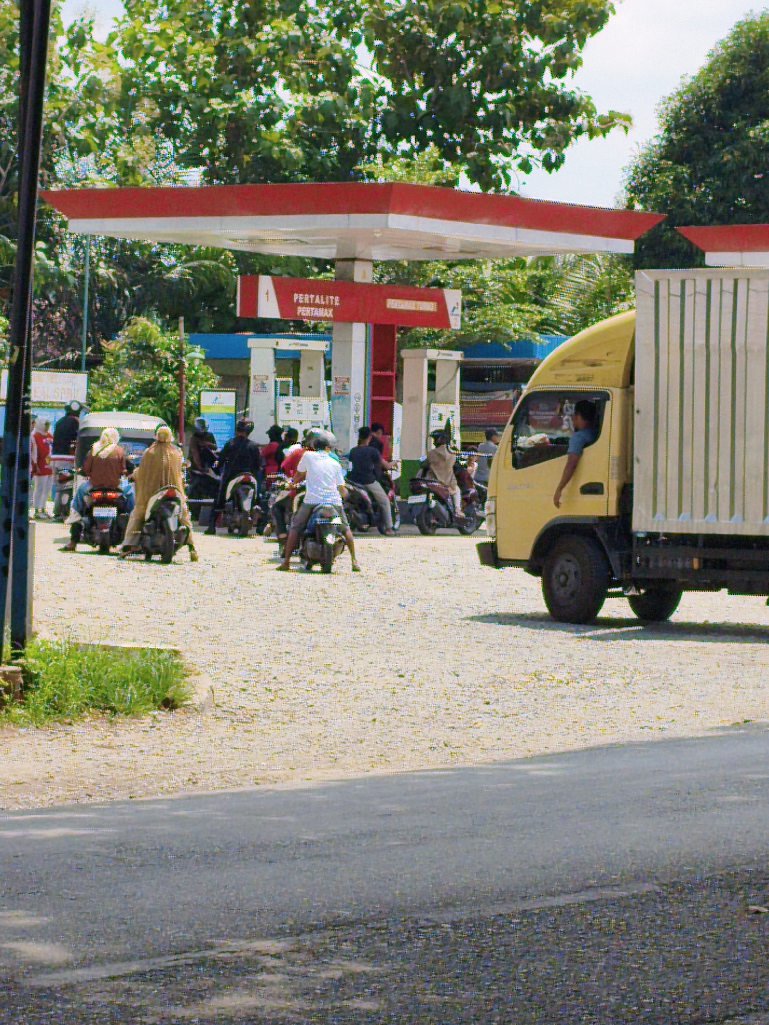 The image depicts an outdoor scene at a Pertamina gas station in the City of Tebing Tinggi, Indonesia, likely during daylight hours. A yellow truck is parked on the right side of the frame, with its driver visible inside, his arm resting on the open window. In the background, a gas station canopy with red and white accents is present, along with fuel pumps. Several people on motorcycles are also visible, some waiting in line, others parked, suggesting a busy period at the station. The ground is covered with gravel and sand, and lush green trees and foliage are abundant in the background, indicating a tropical or subtropical environment. The overall atmosphere appears to be that of a typical day at a roadside service station.