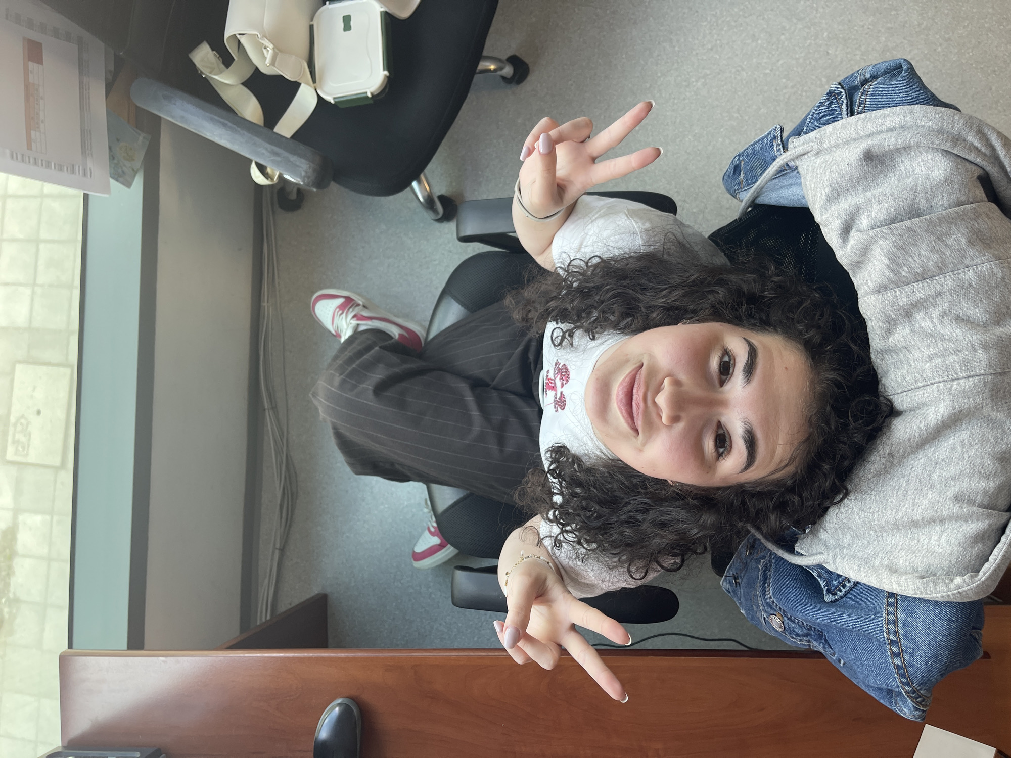 An overhead shot depicts a young person seated in a black office chair within an indoor office or workspace environment. The individual, with dark curly hair, looks up at the camera with a subtle smile and makes a peace sign with both hands.
The person is dressed in a white t-shirt featuring a faint red and white embroidered design, dark pinstriped trousers, and white sneakers with pink accents. Light pink nail polish is visible on their fingernails, and a light-colored bracelet is worn on the left wrist.
To the person's left, another black office chair holds a white rectangular container with a green lid, alongside a beige strap. A large window beyond this reveals an outdoor scene, suggesting daylight. Papers with illegible text and tables rest on a surface near the window. White cables run along a wall in the background.
A grey hoodie and a blue denim jacket are draped over the back of the person's chair to their right. A wooden desk or table occupies the foreground, with a black shoe visible on the floor beside it. The floor is grey. The overall atmosphere conveyed by the person's expression is pleasant and relaxed. There are no specific visual cues in the image to directly indicate the location as Sofia, Bulgaria.