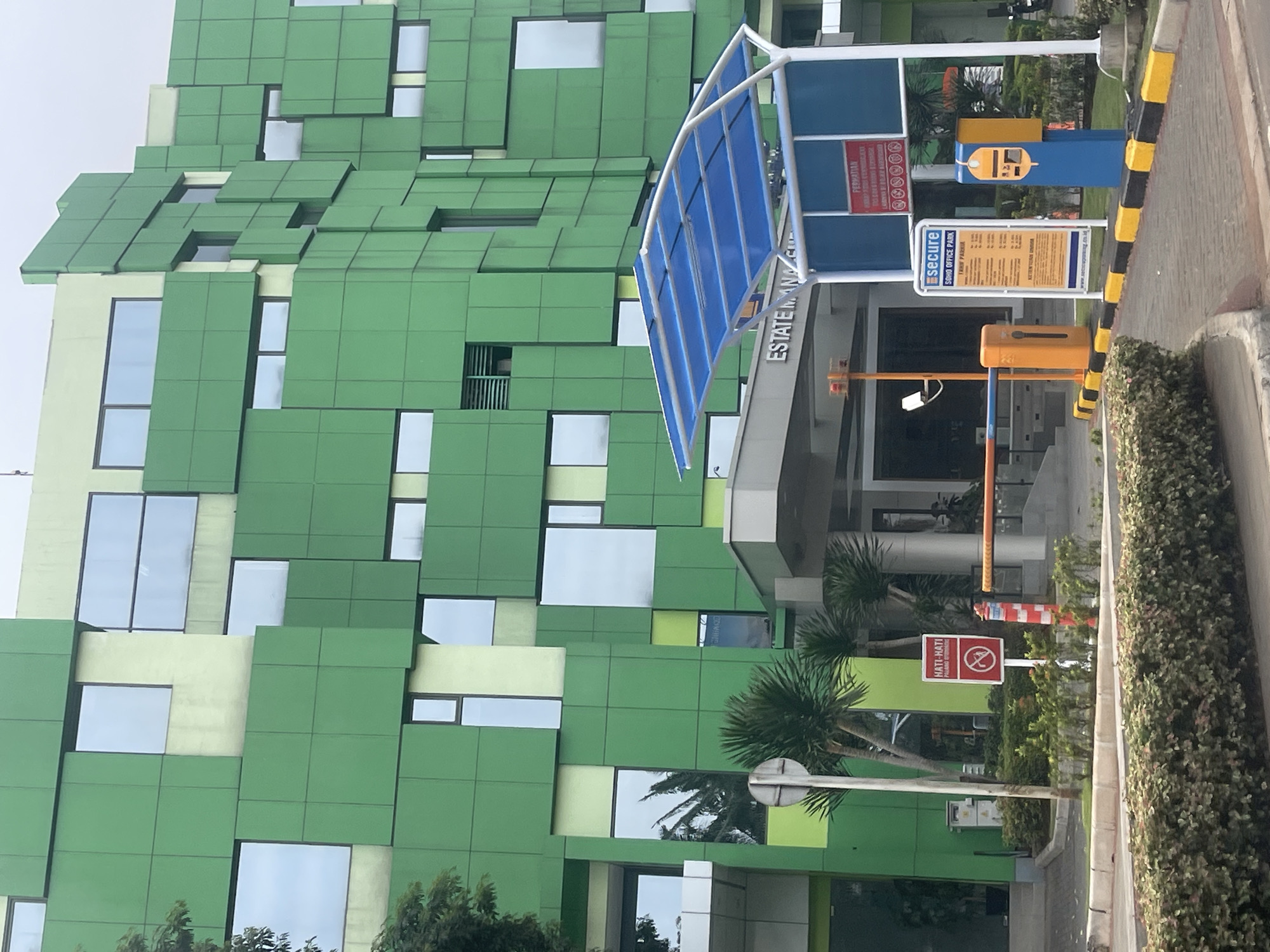 The image shows the exterior of a modern, multi-story building with a distinctive green facade composed of irregular geometric panels and numerous windows of varying sizes. The building is located in Curug Sangereng, Indonesia. In the foreground, there is a paved area with a blue canopy shelter, a parking barrier with an orange dispenser, and a yellow and blue ticket machine. A red and white sign with text and an icon indicating "HATI-HATI" (be careful) is visible on the left side of the image, near some palm trees and bushes. To the right of the barrier, there is another sign indicating "SOHO OFFICE PARK". The overall scene depicts an entrance area to an office park, suggesting a commercial or business district.