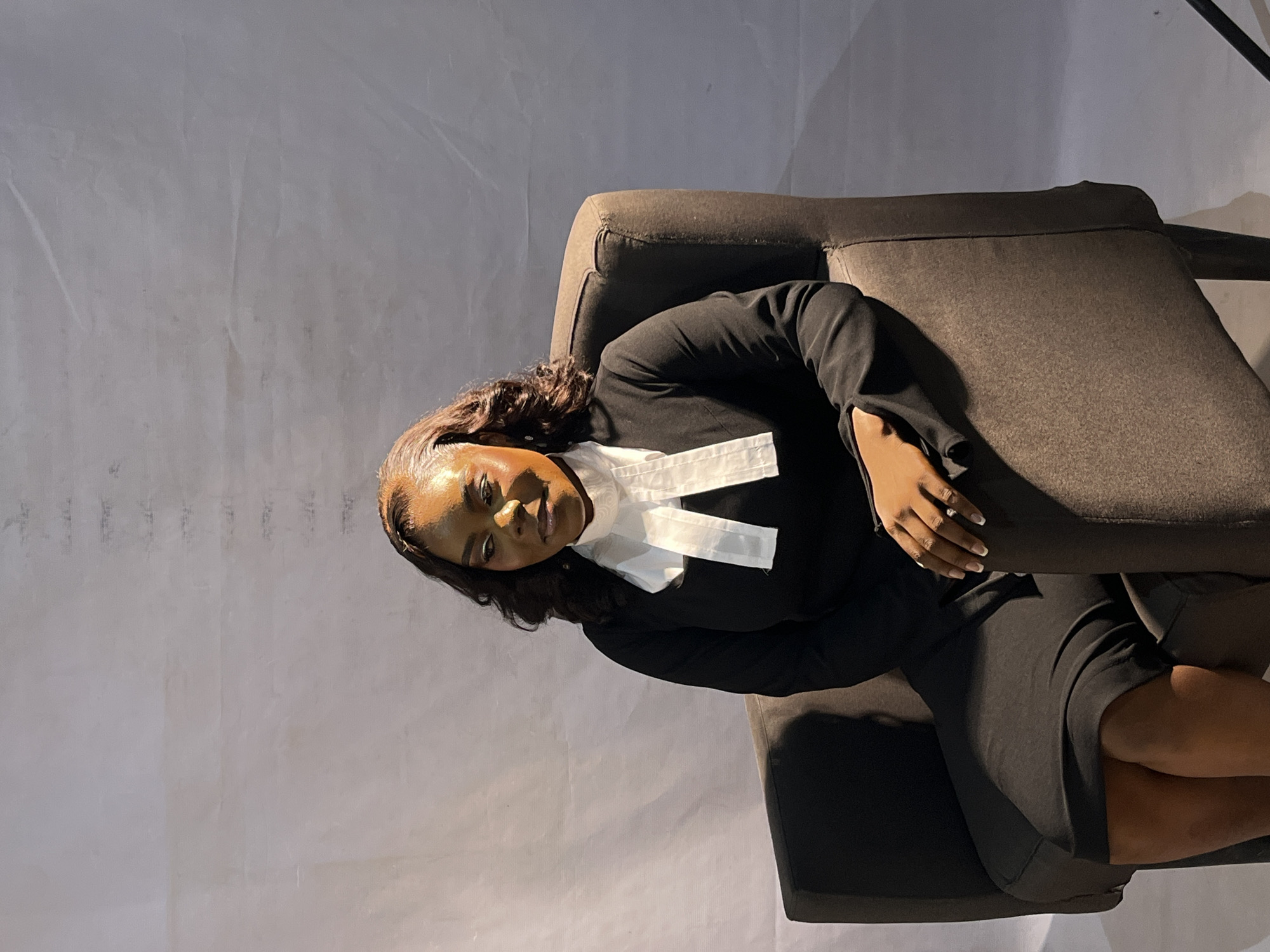 A woman is seated in a dark gray armchair against a plain, textured gray background. She is wearing a black dress with white clerical bands around her neck. Her hair is dark brown and styled in loose curls. She is looking to her left, with her right hand resting on her lap. The lighting is soft, with shadows cast to her right. The overall scene is a studio portrait.