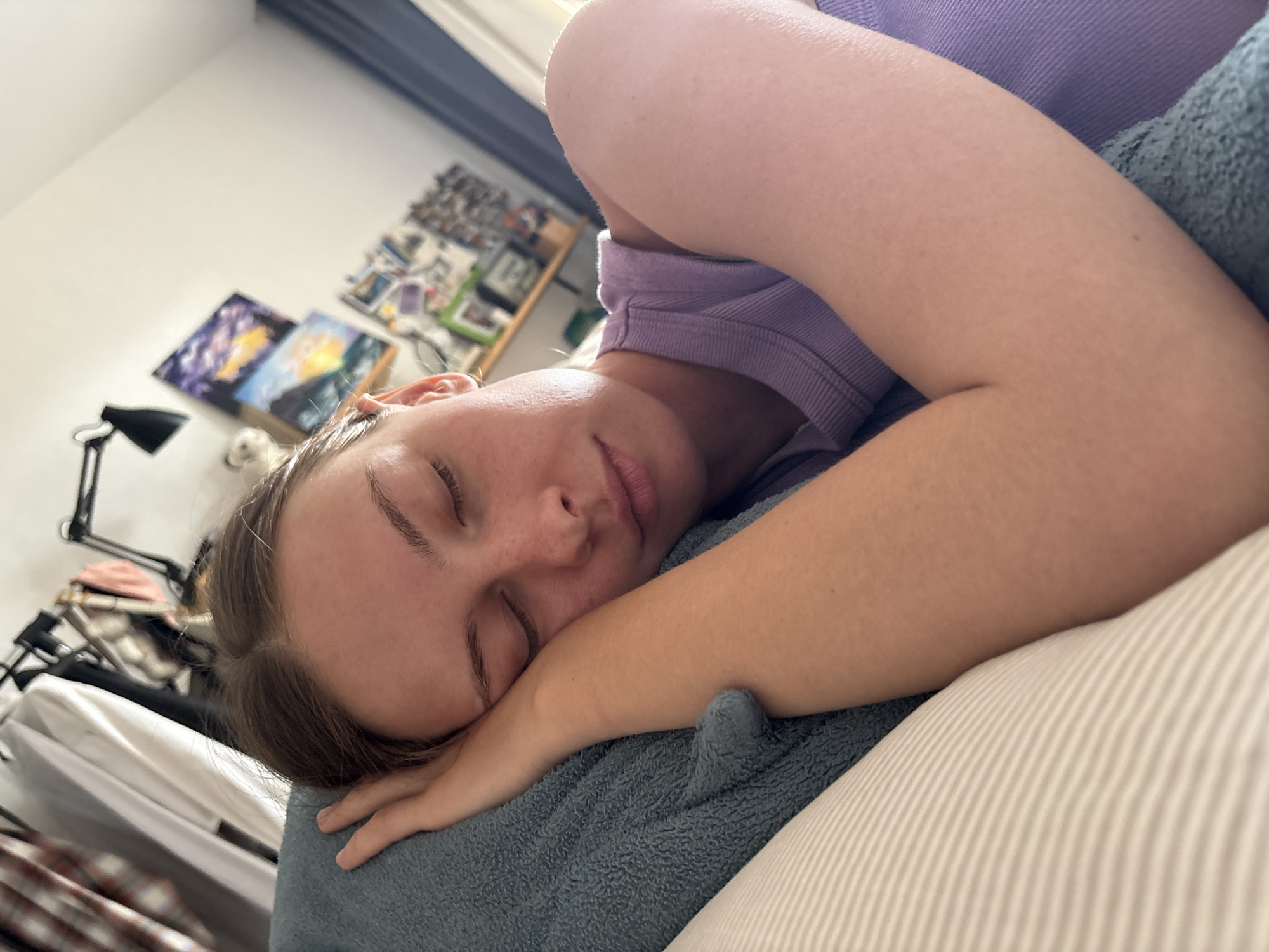 A young adult female is observed sleeping or deeply resting on a pillow in an indoor setting, situated in the city of Perm, Russia. She lies on her right side, with her head on a white pillow featuring subtle light grey stripes, and her left arm bent, resting on a blue, textured blanket or towel. Her eyes are closed, and her facial expression is relaxed. She has light brown hair, parted centrally, and is wearing a light purple ribbed tank top. The background on the left reveals a clothes rack with various garments hanging. Above and to the right, a black adjustable desk lamp is visible against a white wall. Below the lamp, two framed landscape paintings are displayed, one with a purple-toned sky and another depicting a sunset over water. Further right, a collection of smaller framed pictures or postcards is arranged, and a dark blue curtain is partially visible along the far right. The room is illuminated by natural light, indicating daytime.