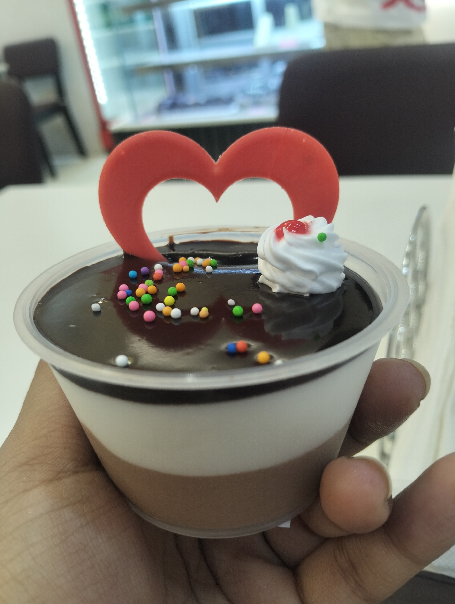 A hand is holding a plastic cup of dessert with multiple layers. The bottom layer appears to be chocolate mousse. The next layer is a white mousse. The top layer is a shiny chocolate ganache with colorful sprinkles and a dollop of whipped cream with red and green decorations. A red heart-shaped decoration is stuck in the top of the dessert. The background is blurred, but shows a light-colored table and chairs. The setting is likely a dessert shop in Dhaka, Bangladesh.