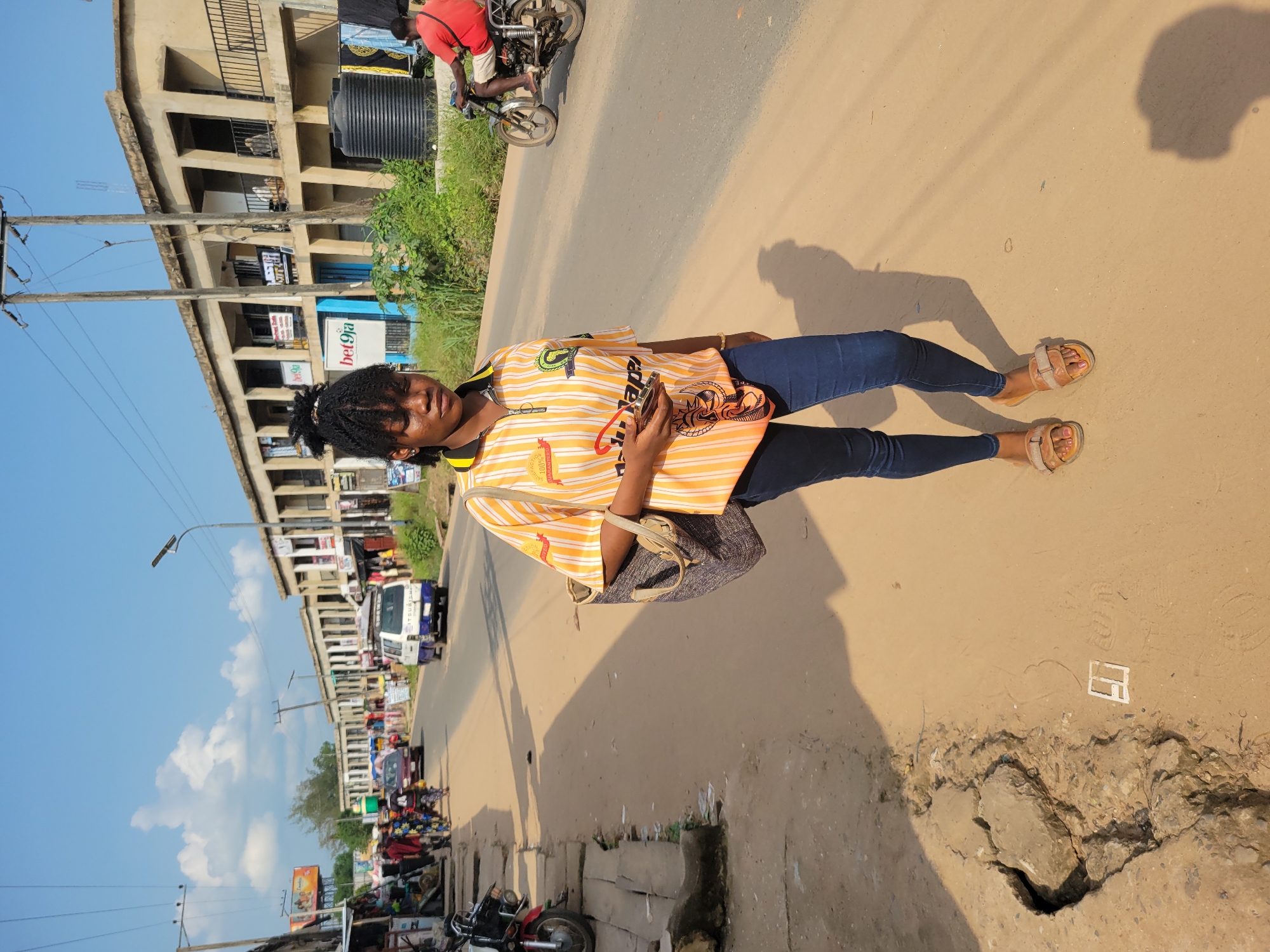 A young woman stands on a dusty road in Egbagu, Nigeria. She is wearing a yellow and orange striped t-shirt and dark blue jeans, with tan sandals on her feet. She carries a dark patterned bag over her shoulder. Her hair is styled in braids, and she has a calm expression on her face. In the background, there are buildings with balconies and an assortment of shops lining the street. A white truck with "MITSUBISHI" visible on its side is parked on the road, along with other vehicles and motorcycles. People can be seen milling about the shops and on the street, suggesting a busy marketplace or commercial area. Overhead, a bright blue sky with scattered white clouds indicates a clear day. The sun is casting strong shadows, suggesting it is likely midday or early afternoon. A large black water tank is visible on top of one of the buildings. Signs for "bet9ja" are also visible on a building. The overall scene depicts a vibrant street in a Nigerian town.