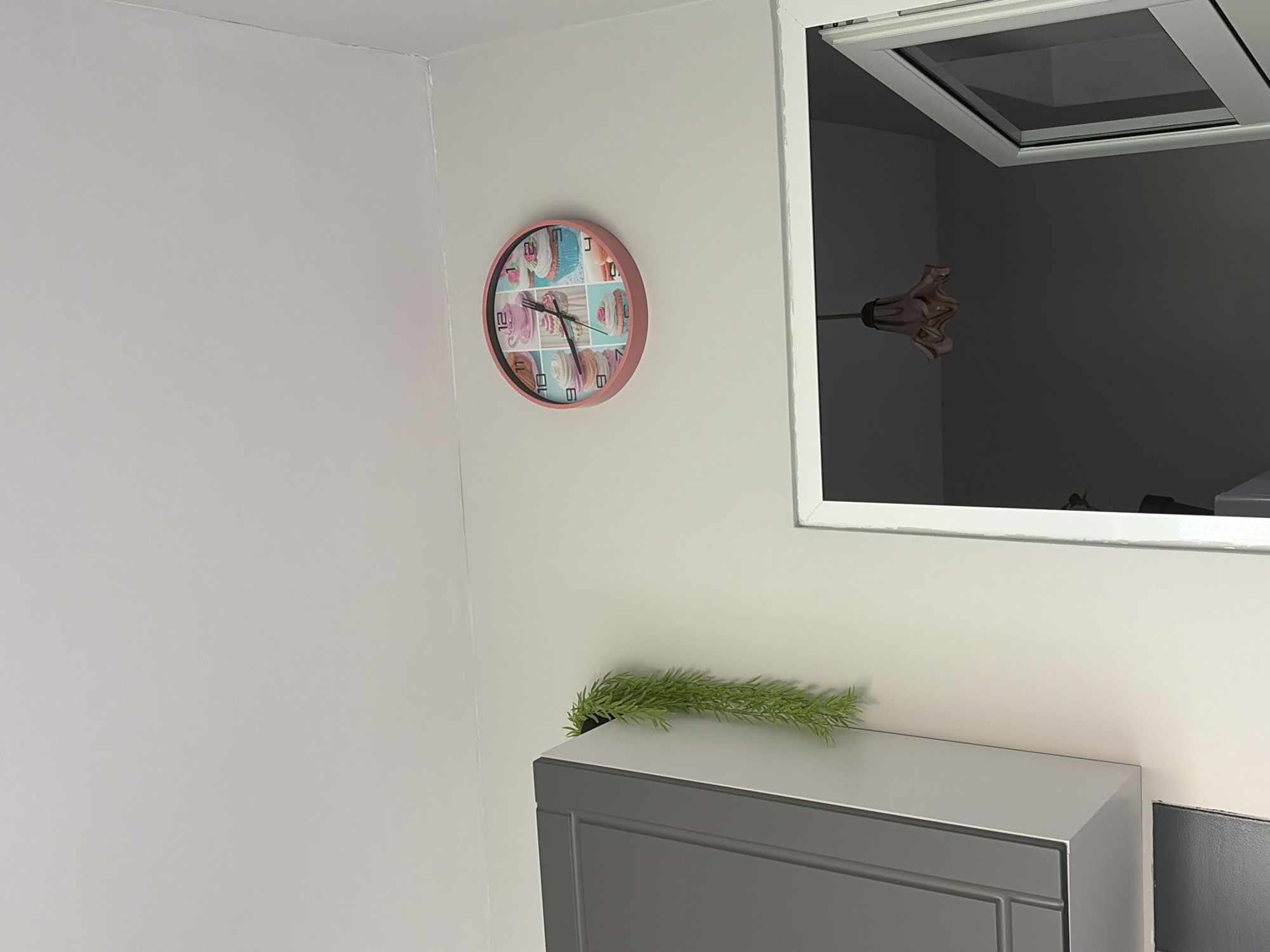 This image presents an interior scene, likely a room within a modern home in Stara Zagora, Bulgaria. The walls are painted a light, possibly off-white or very light grey, creating a clean and bright aesthetic.
A round wall clock with a pink or rose gold frame is centrally placed on the wall. Its face features a grid of nine squares, each depicting a different colorful cupcake or pastry in soft pastel tones. The clock's black hour and minute hands indicate the time as approximately 12:45. The numbers 12, 10, 9, and 8 are clearly visible on the clock face.
To the right, a white-framed opening, possibly a window or a recessed niche, reveals a dark, shadowed interior space beyond. Within this dark area, a reflection of what appears to be a decorative light fixture or object is visible. It has a multi-lobed, organic shape and is colored light brown or pinkish, attached to a slender rod.
Below this opening, a grey cabinet or storage unit with a flat top surface and a paneled front sits against the wall. A long, slender artificial green plant or foliage is draped casually on top of the cabinet, adding a touch of natural accent to the setting.
The scene is static, with no people visible and no specific activity taking place. The overall impression is one of a well-maintained, tidy space, likely captured during daytime given the general lighting.