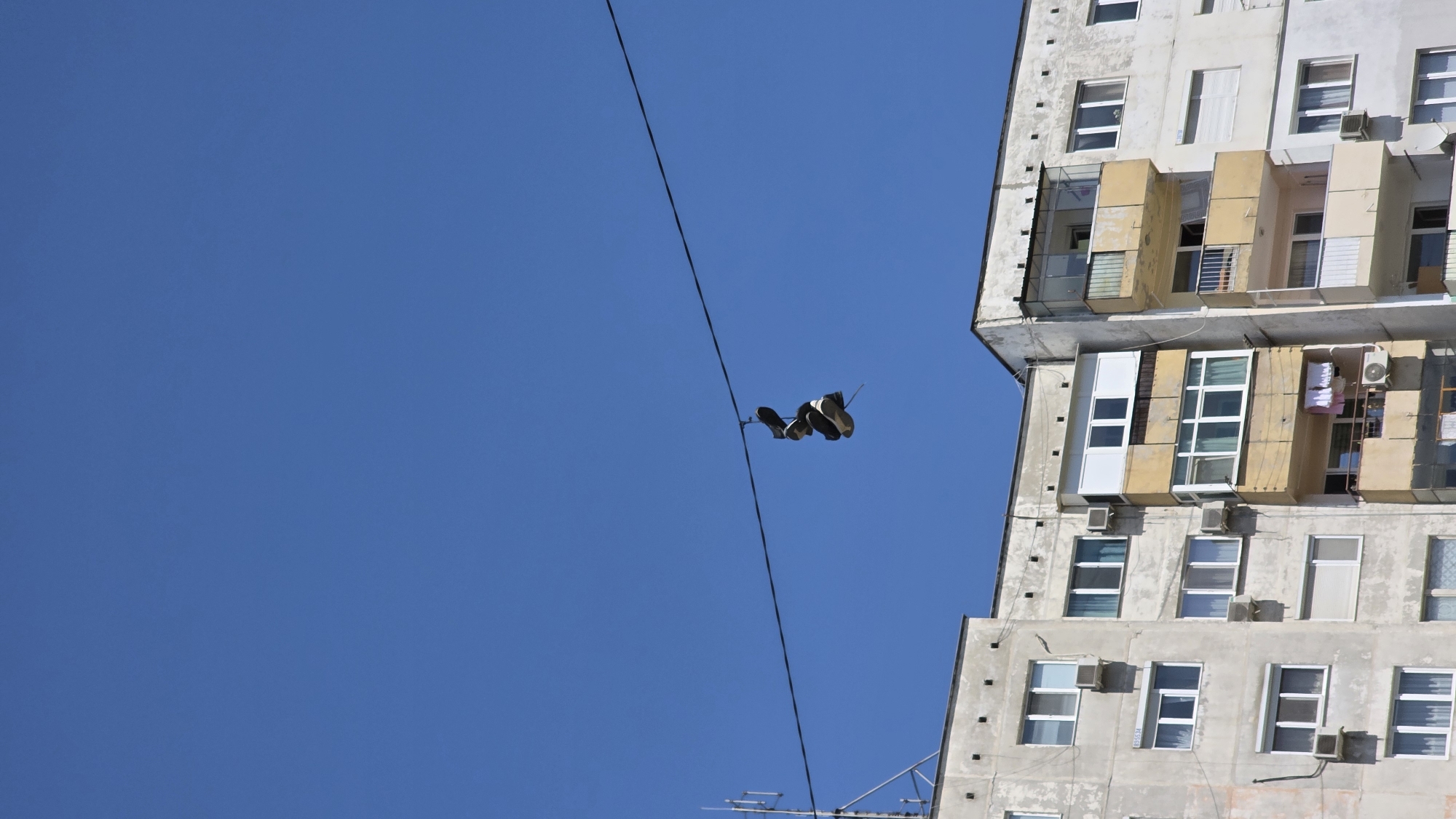 This low-angle, outdoor shot captures a vibrant urban scene in Blagoevgrad, Bulgaria, under a clear, bright blue sky on a sunny day. The dominant features are a tall, multi-story building on the right side of the frame and a pair of shoes suspended from a wire against the expansive sky.
The building, likely an apartment block, is constructed of light-colored concrete, showing some signs of wear and various repairs. It features numerous windows, many with blinds or curtains visible, and a variety of small balconies. Some balconies are enclosed with glass or simple railings, while others appear more open, adorned with personal items or external air conditioning units. The architectural style suggests a typical Eastern European residential structure.
Stretching diagonally across the vast blue sky is a single, dark cable, presumably an electrical or telecommunications line. From this wire hangs a pair of dark-colored shoes, appearing to be sneakers or trainers, suspended by their laces. There is no visible activity beyond the static scene of the hanging shoes and the building. No text is discernible in the image.