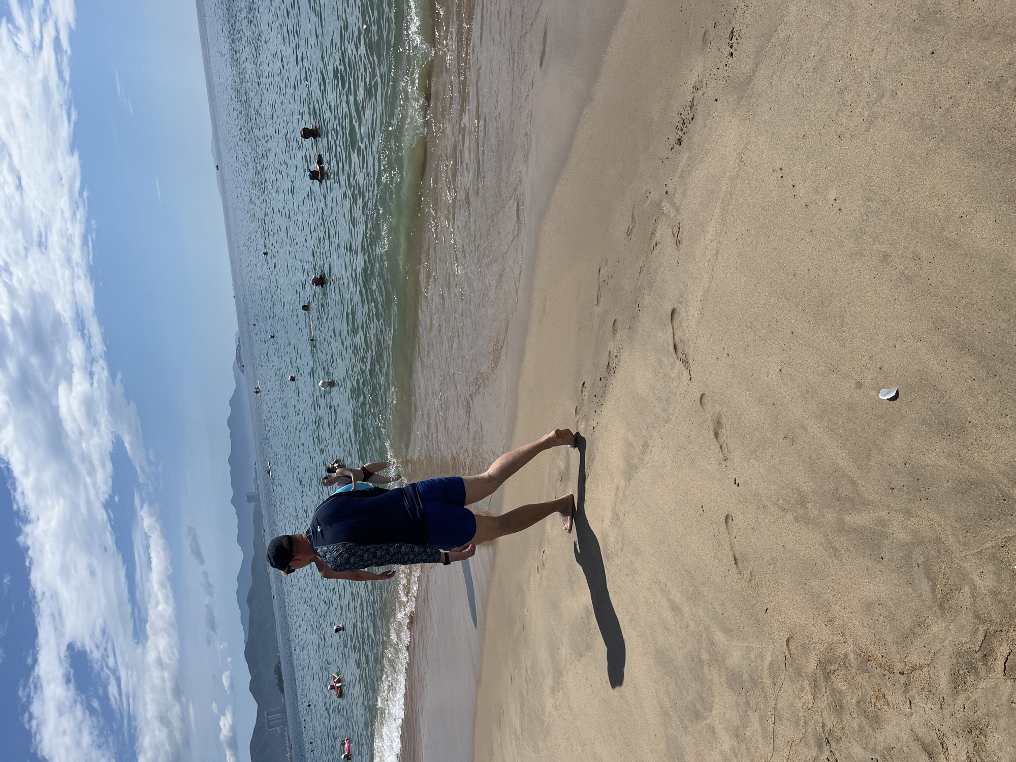 This image captures a sunny day on a sandy beach in Nha Trang, Vietnam, with the ocean in the background. A person in a dark blue rashguard and shorts is walking away from the camera on the wet sand near the water's edge, casting a shadow. In the shallow water, several other individuals are visible, some swimming and others standing. A few pink inflatable rings are also present in the sea. The shoreline is lined with a gentle surf. The horizon features a hazy mountain range and a clear blue sky with scattered clouds. The sand shows footprints, indicating recent activity.