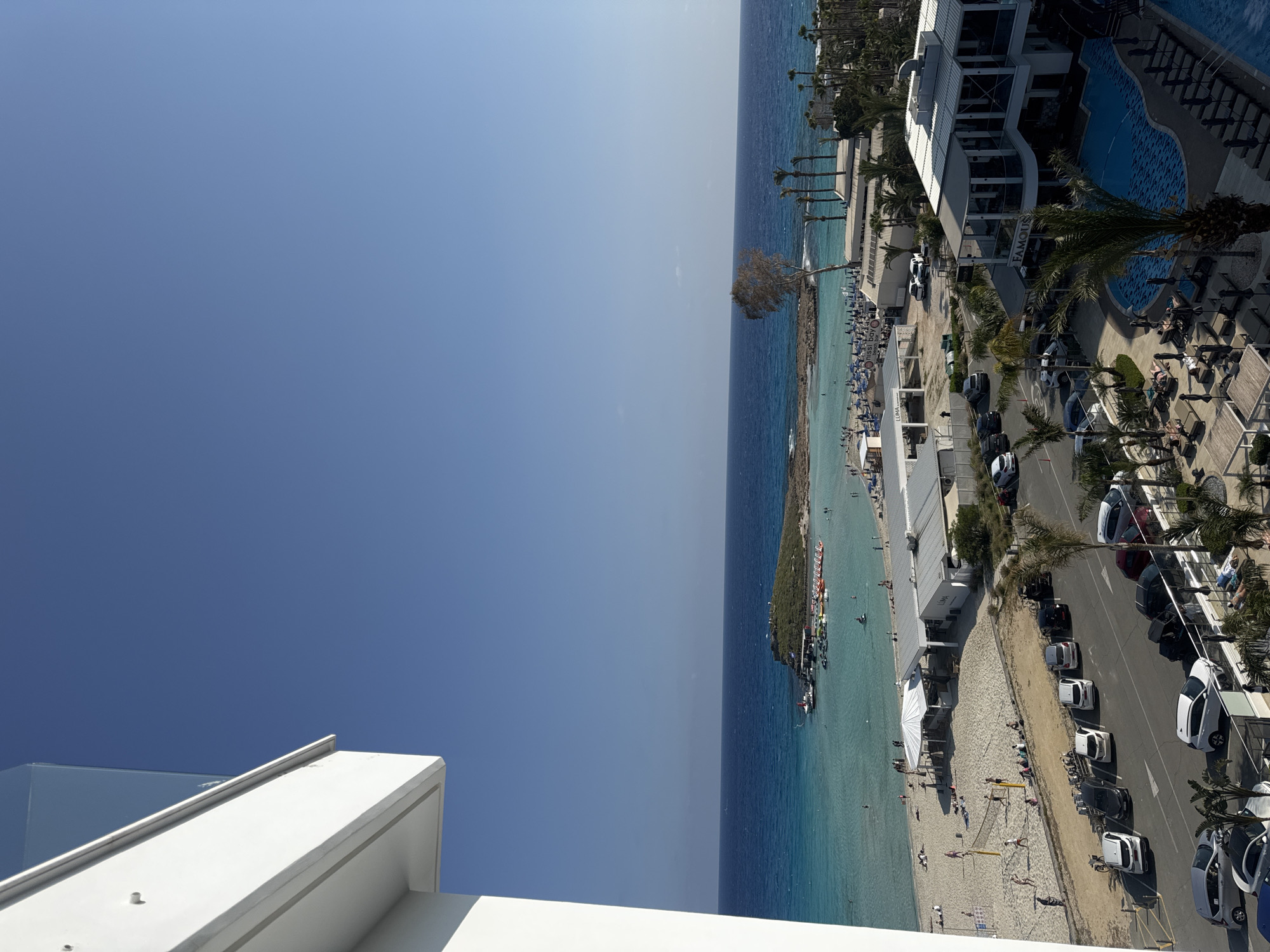 This image presents a vibrant, sun-drenched coastal scene, likely captured from an elevated position overlooking a bustling beach resort in Ayia Napa, Cyprus. The bright, clear blue sky dominates the upper portion, meeting the horizon where the deep blue sea begins.
The coastline features a long stretch of sandy beach, populated by numerous people enjoying the sunny weather. Many individuals are seen lounging on sunbeds under umbrellas, walking along the shore, or wading and swimming in the calm, turquoise waters. A volleyball net is set up on the sand, indicating recreational activity. Further out in the sea, a small, verdant island or peninsula is visible, surrounded by boats and water sports equipment, suggesting water-based activities. A solitary, leafless tree stands out near the water's edge on the main beach.
Parallel to the beach runs a paved road, where a variety of cars are parked. On the inland side of the road, several modern, white resort buildings line the street. One prominent building has "FAMOUS" written on its facade and features a swimming pool with a patterned bottom. Another building further along the coast has "LUMA" visible on its sign. Palm trees are scattered throughout the resort area, enhancing the tropical ambiance. People are also seen walking on sidewalks and around the buildings.
The time of day appears to be mid-morning or early afternoon, given the bright sunlight and clear shadows. The weather is perfectly sunny with no clouds, creating an inviting atmosphere for leisure and holiday activities. The scene clearly depicts a lively and popular tourist destination.
