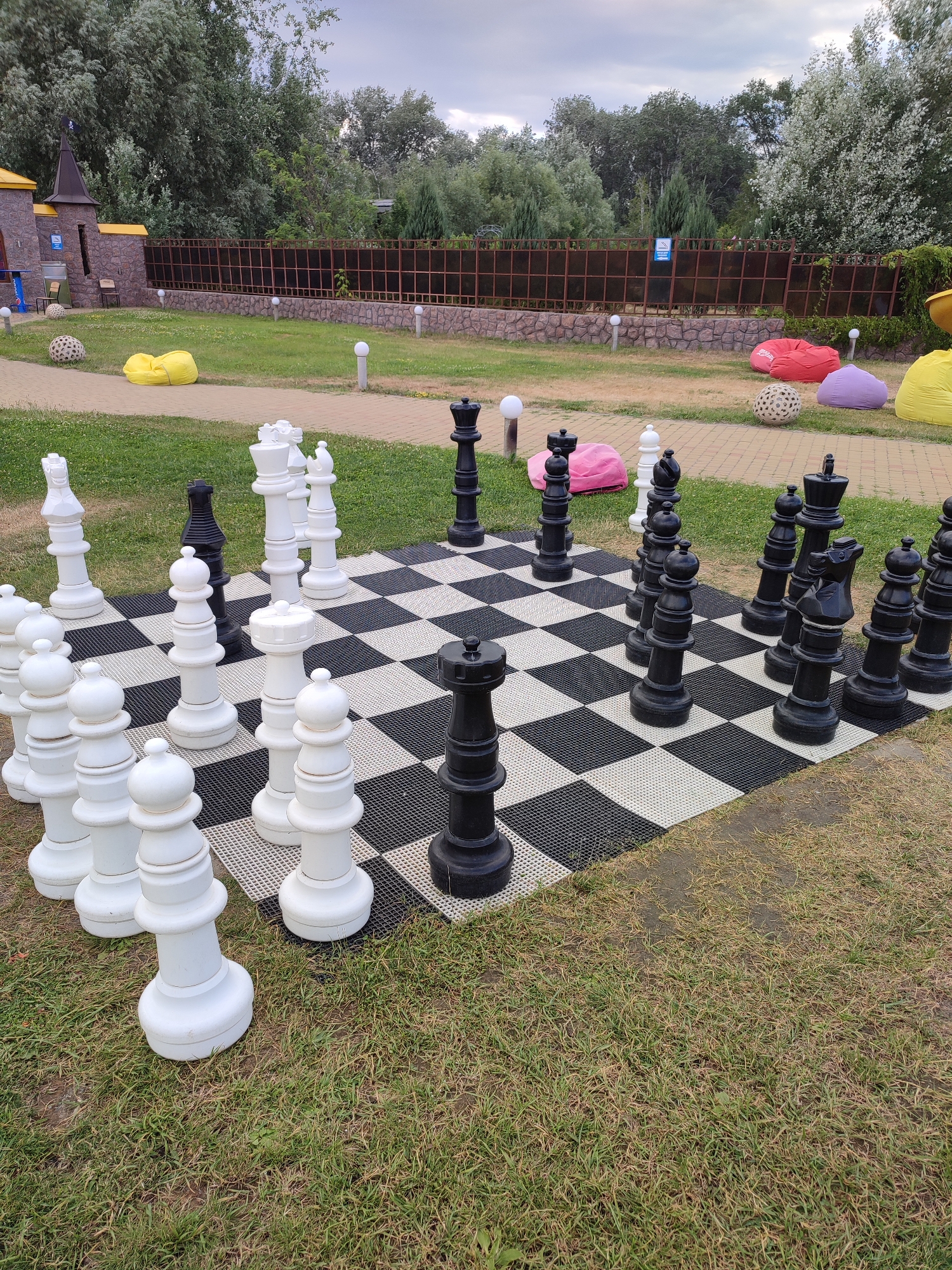 The image shows a life-sized outdoor chess set on a checkered mat. The chess pieces are predominantly white and black, with several white pawns and a white queen in the foreground. Behind them, a black king and queen are visible, along with other black pieces. The chess set is placed on a grassy area, with a paved walkway visible in the background. Beyond the walkway, there is a low stone wall topped with a dark metal fence. In the distance, there are trees and a cloudy sky. To the left of the chess set, a small, castle-like structure with yellow turrets is partially visible. Scattered around the scene are several large, colorful beanbag chairs in yellow, pink, and purple, as well as decorative spherical objects. The overall setting appears to be a recreational area, possibly a park or a resort, in Shcherbani, Ukraine. The time of day seems to be daytime, with overcast weather indicated by the sky. No people are visible in the image.