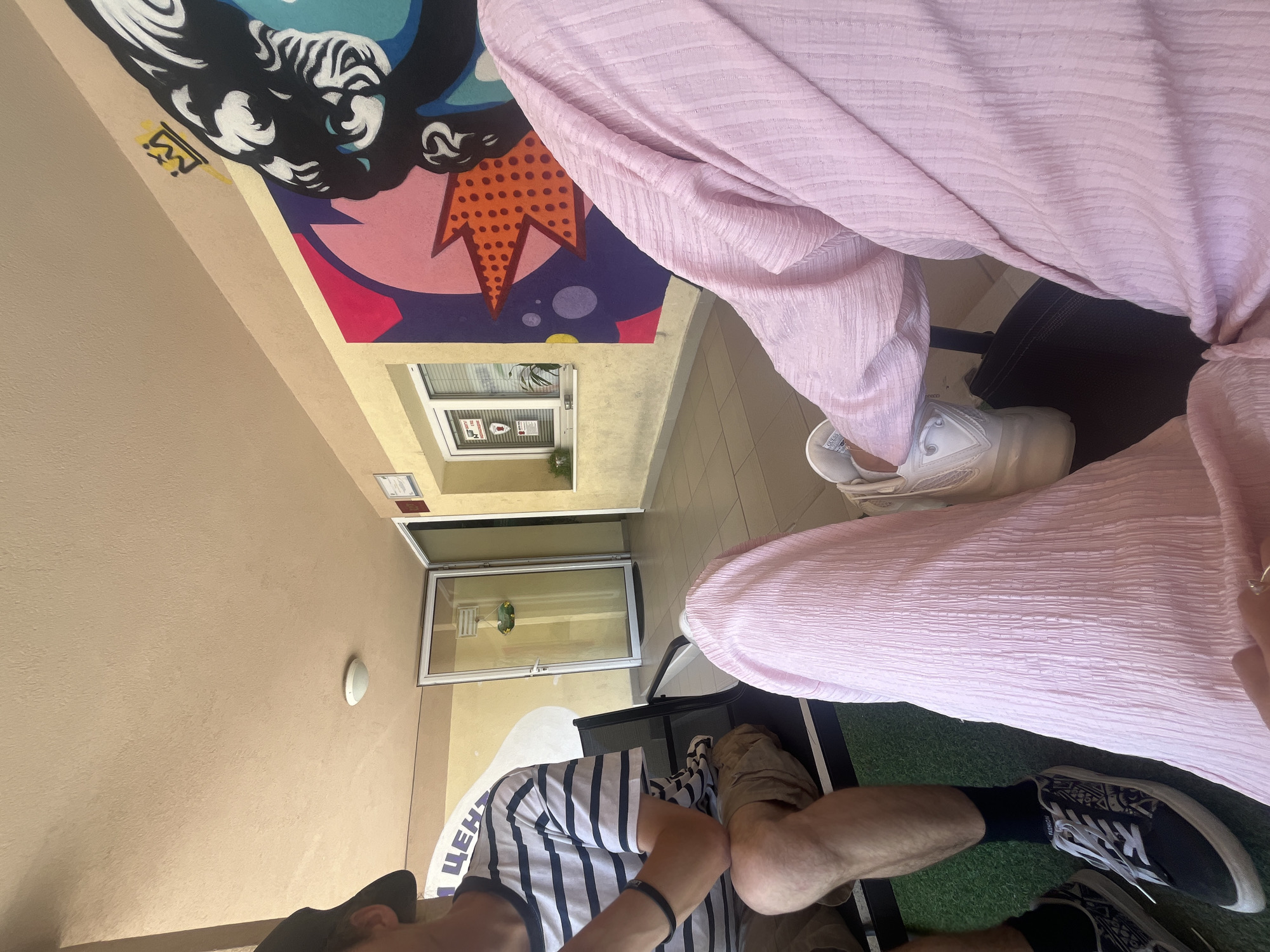 This image depicts two individuals in a waiting area, likely a clinic or a public office, located in Vratsa, Bulgaria. One individual, seen from the chest down, is wearing a pink textured garment and white sneakers. The other individual, partially visible on the left, is wearing a striped t-shirt, shorts, and black sneakers. The environment features light-colored walls, a tiled floor, and a glass door leading to an interior space. A vibrant mural adorns the wall, featuring abstract shapes and a stylized portrait. The lighting suggests it is daytime.
