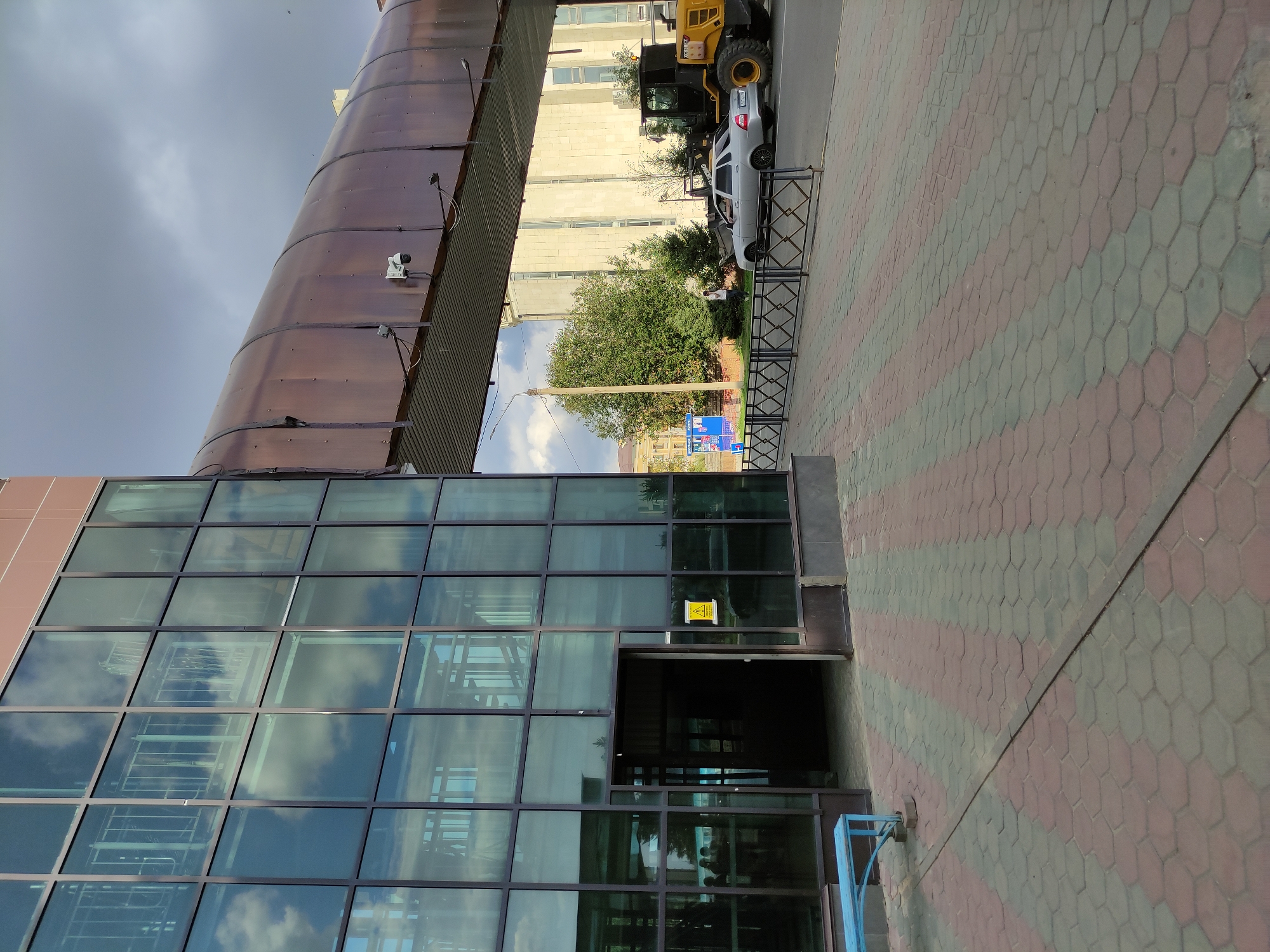 The image depicts an outdoor scene in the city of Semey, Kazakhstan, featuring a modern building with a glass facade and a covered pedestrian walkway. The building's exterior is a striking combination of dark-framed glass panels reflecting a cloudy sky, with sections of reddish-brown material. An open doorway on the ground floor reveals a dark interior, possibly a lobby or entrance. In front of the building, a blue metal bench sits on a patterned sidewalk made of hexagonal concrete pavers in red and gray.
Extending from the building is a brown, semi-transparent covered walkway, supported by metal structures. Visible on the underside of the walkway are security cameras and lighting fixtures. Beyond the walkway, a street is visible with a silver sedan parked and a yellow construction vehicle, possibly a front-end loader, further down the road. Trees and other buildings form the backdrop. The overall impression is one of an urban environment with commercial or public infrastructure.