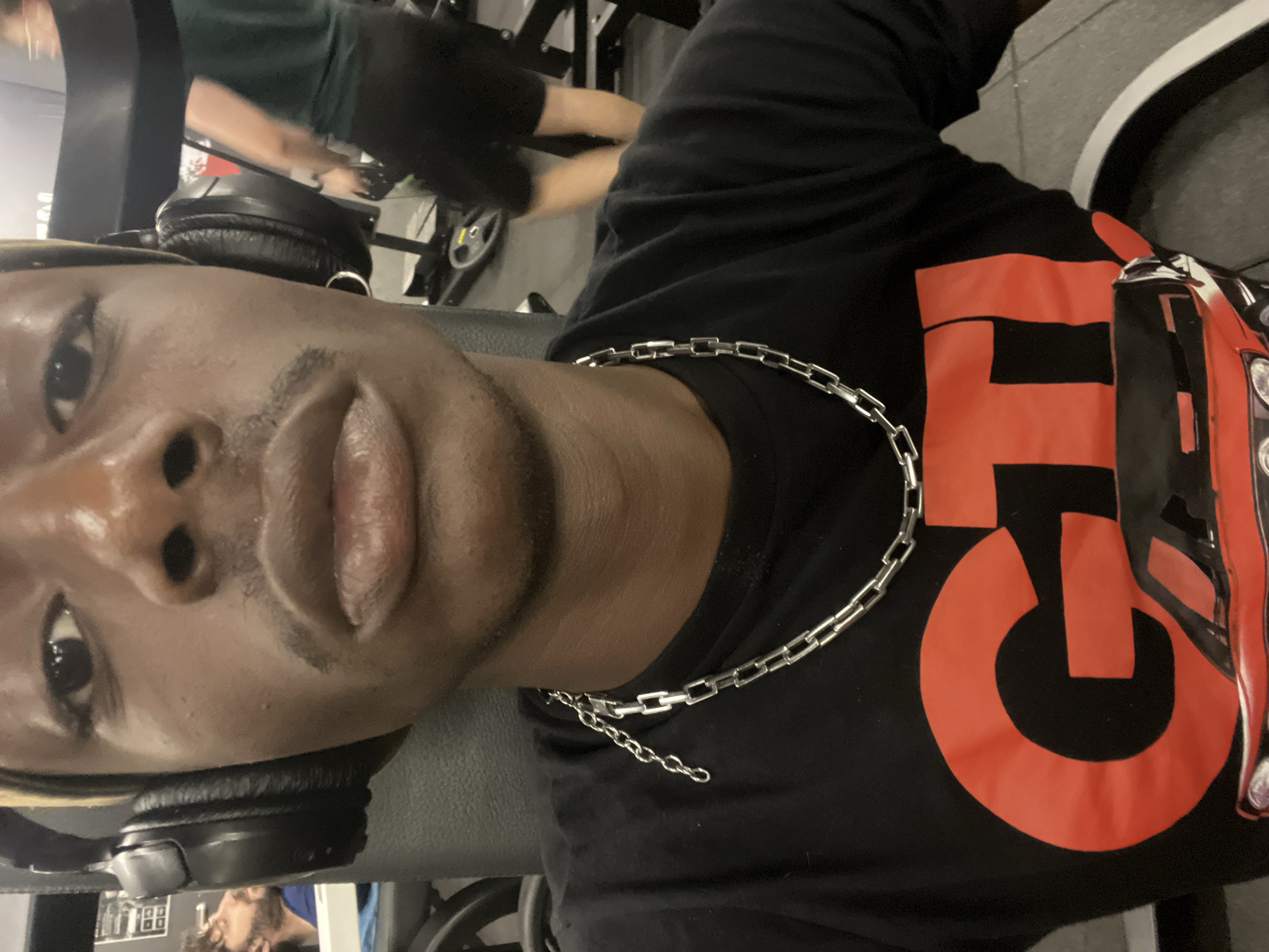 A close-up, tilted-angle photograph depicts a man, presumably in a gym setting. He is wearing headphones around his neck and a silver chain. His black t-shirt features a prominent red "GTI" design. The background shows blurred gym equipment and another individual exercising. The image lacks discernible details to confidently establish a location in Barcelona. Therefore, that part of the request cannot be fulfilled.