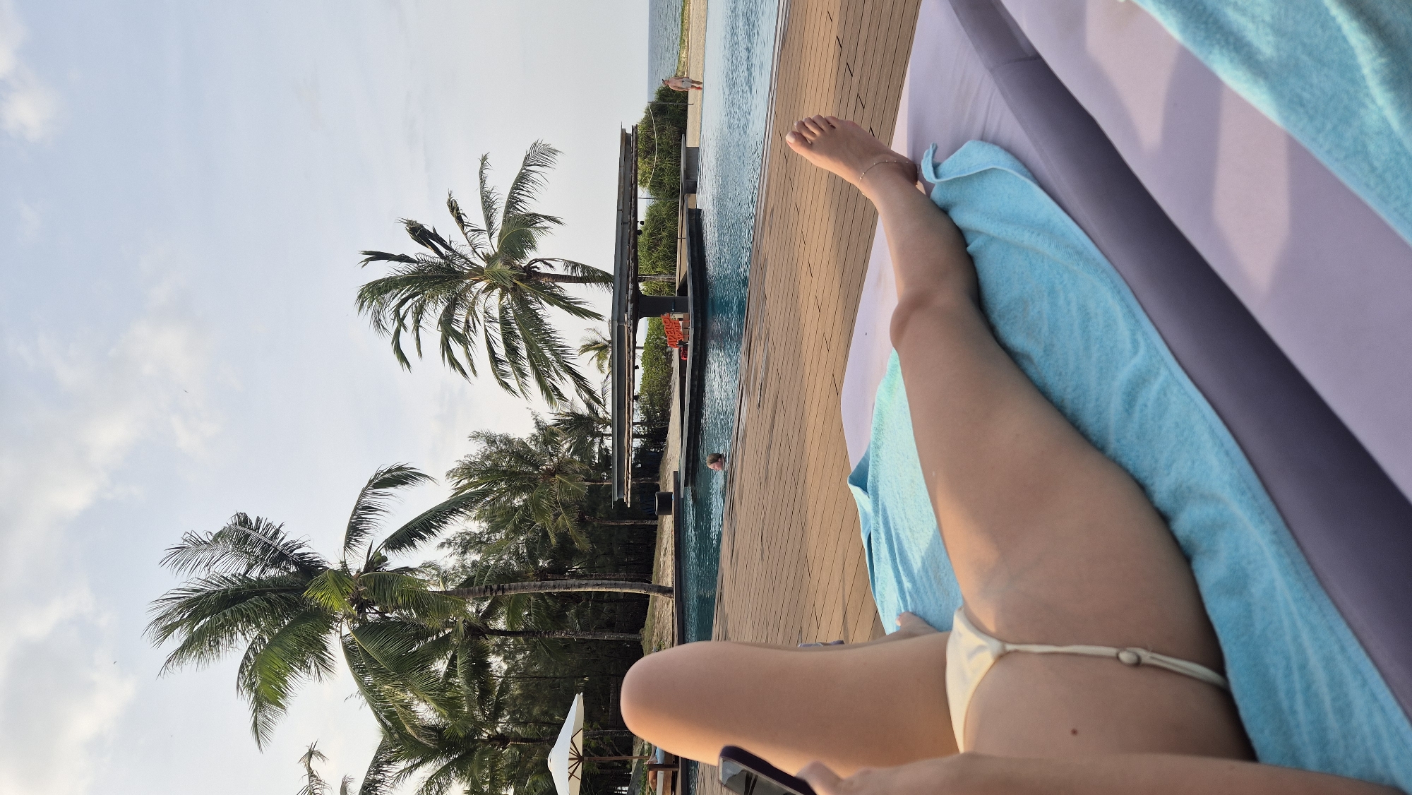 The image shows a person's legs and torso, lying on a lounge chair covered by a blue towel, with a person in the background swimming in a pool. The background features palm trees, a pool bar, and the ocean. The location is a resort in Hoi An, Vietnam, with the ocean and beach visible in the distance. The sky is partly cloudy.