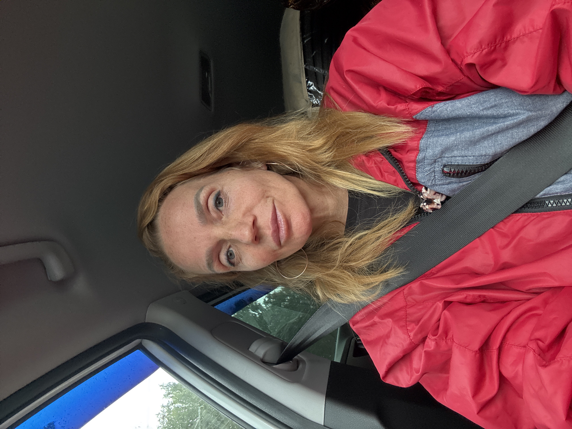 A candid, medium shot shows a woman with blonde hair and light-colored eyes looking directly at the camera, seated inside a vehicle. She is wearing a red and gray jacket with a black t-shirt visible underneath, and a black seatbelt is fastened across her chest. The woman is wearing hoop earrings and has makeup on her face, including lip gloss. The interior of the car is visible, with a gray headliner and a blue tinted window on the left side of the frame. The background outside the window shows trees and foliage, suggesting an outdoor, possibly rural or park-like setting. The scene is overcast, with rain visible on the window. The location within Saint Petersburg, Russia is not specified.