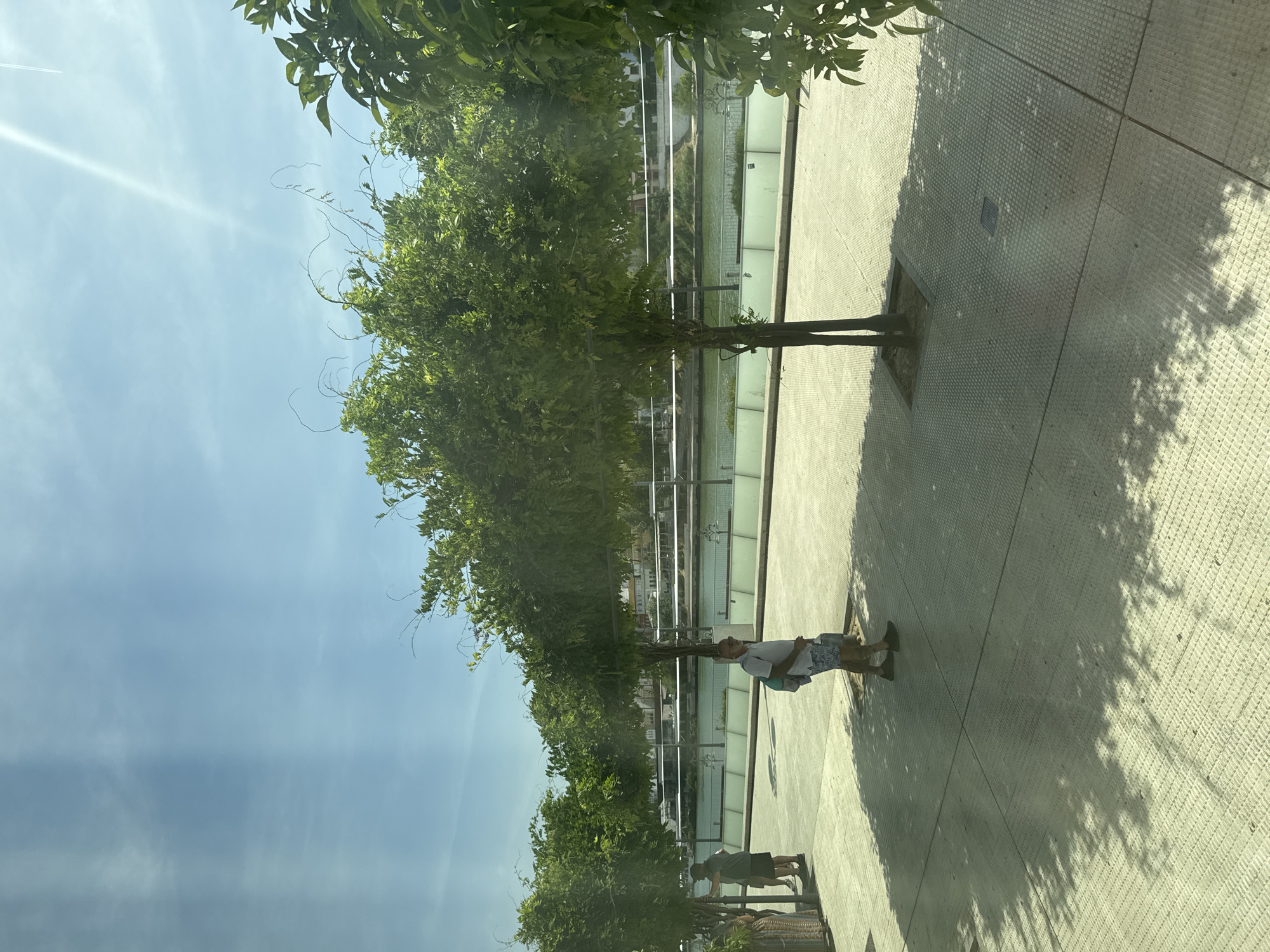 The image captures a sunny day outdoors, likely along a waterfront in Seville, Spain, given the architectural style of buildings visible across the river and the presence of lush greenery. The foreground is a paved area with a grid pattern, with several trees casting dappled shadows. In the middle ground, a man with sunglasses, wearing a white t-shirt and patterned shorts, walks towards the right, carrying a blue backpack. Another person, wearing a dark t-shirt and shorts, walks towards the left, with a basket-like object on their left side. A body of water, possibly a river, is visible behind a low wall and railing. Across the water, a line of buildings with light-colored facades and red-tiled roofs can be seen. The sky is blue with some scattered clouds and a bright sun creating lens flare effects.