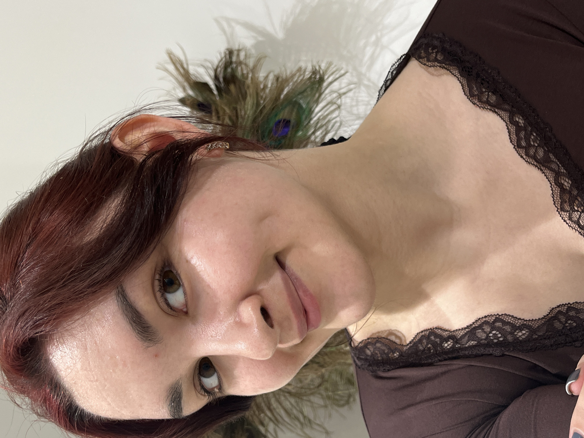 An image taken in Neftekamsk, Russia, depicts a young adult female from the chest up, positioned slightly to the right of the frame's center. She possesses fair skin, dark auburn hair parted on the left, and brown eyes. A subtle, closed-lip smile reveals dimples on both cheeks, and her gaze is directed towards the viewer. A small, red blemish is visible on her forehead. She is wearing a dark brown long-sleeved top featuring black lace trim along the neckline. A small, patterned gold-colored hoop earring is visible in her right ear. Behind her right ear, a cluster of green and blue iridescent peacock feathers is arranged. The background consists of a plain, light-colored wall. A sliver of a hand with dark nail polish is visible in the lower left corner.