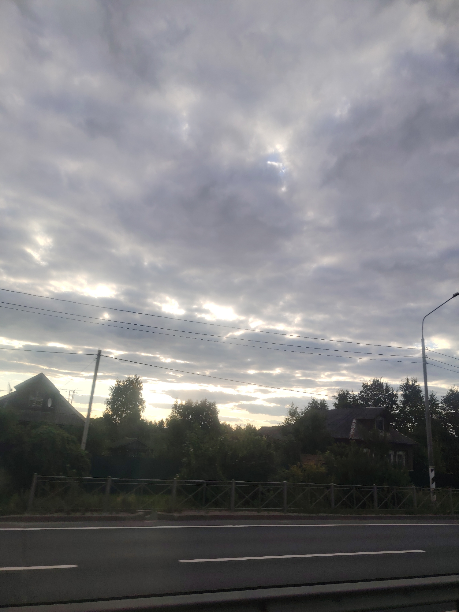 The image shows a roadside view in Golovkovo, Russia, likely during late afternoon or early evening based on the lighting. The sky is overcast with dramatic, layered clouds, through which sunlight is breaking in places, creating bright horizontal bands of light. In the foreground, a dark asphalt road with white lane markings stretches across the bottom of the frame. A metal fence runs parallel to the road, separating it from a grassy area and a line of trees. Behind the fence and trees, several rustic wooden houses with dark roofs are visible, nestled among dense greenery. A streetlamp stands on the right side of the image, and power lines crisscross the sky. The overall atmosphere is muted and somewhat serene, characteristic of a rural or semi-rural setting.