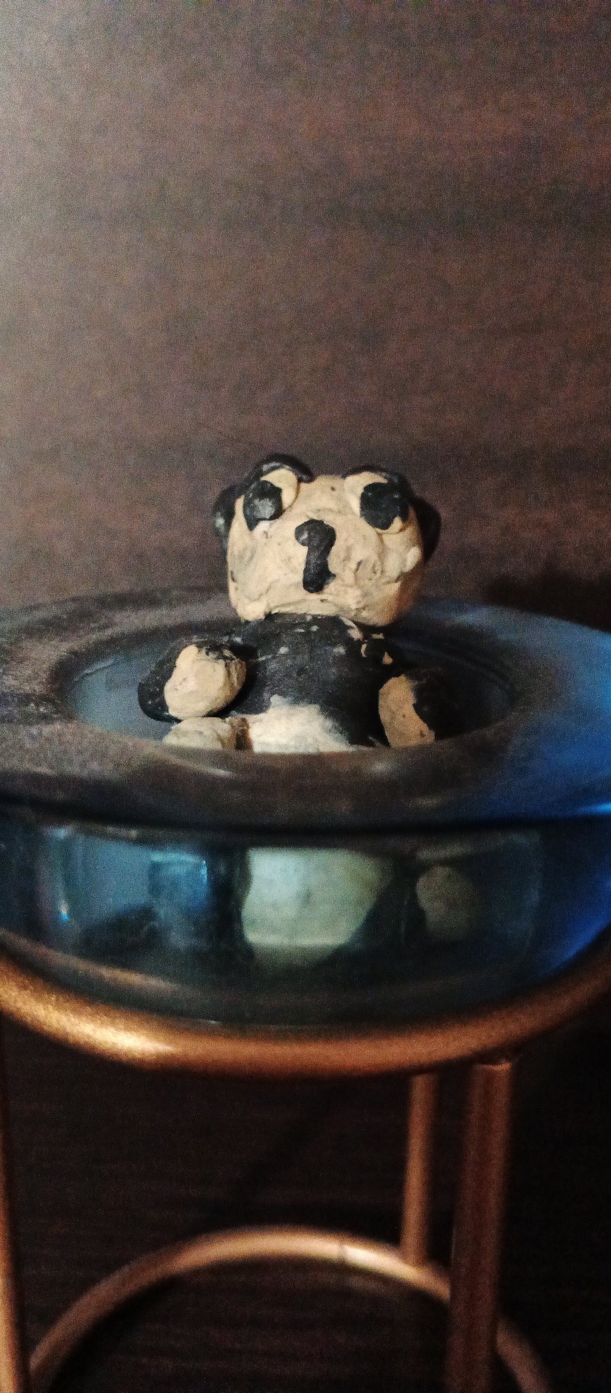 A handmade clay figurine of a black and white bear is positioned in a glass bowl. The figurine has black eye patches and a black nose. The bowl appears to be filled with a clear liquid, and the bowl rests on a stand with bronze-colored legs. The background is a dark, textured surface. The image was taken in Berehove, Ukraine.