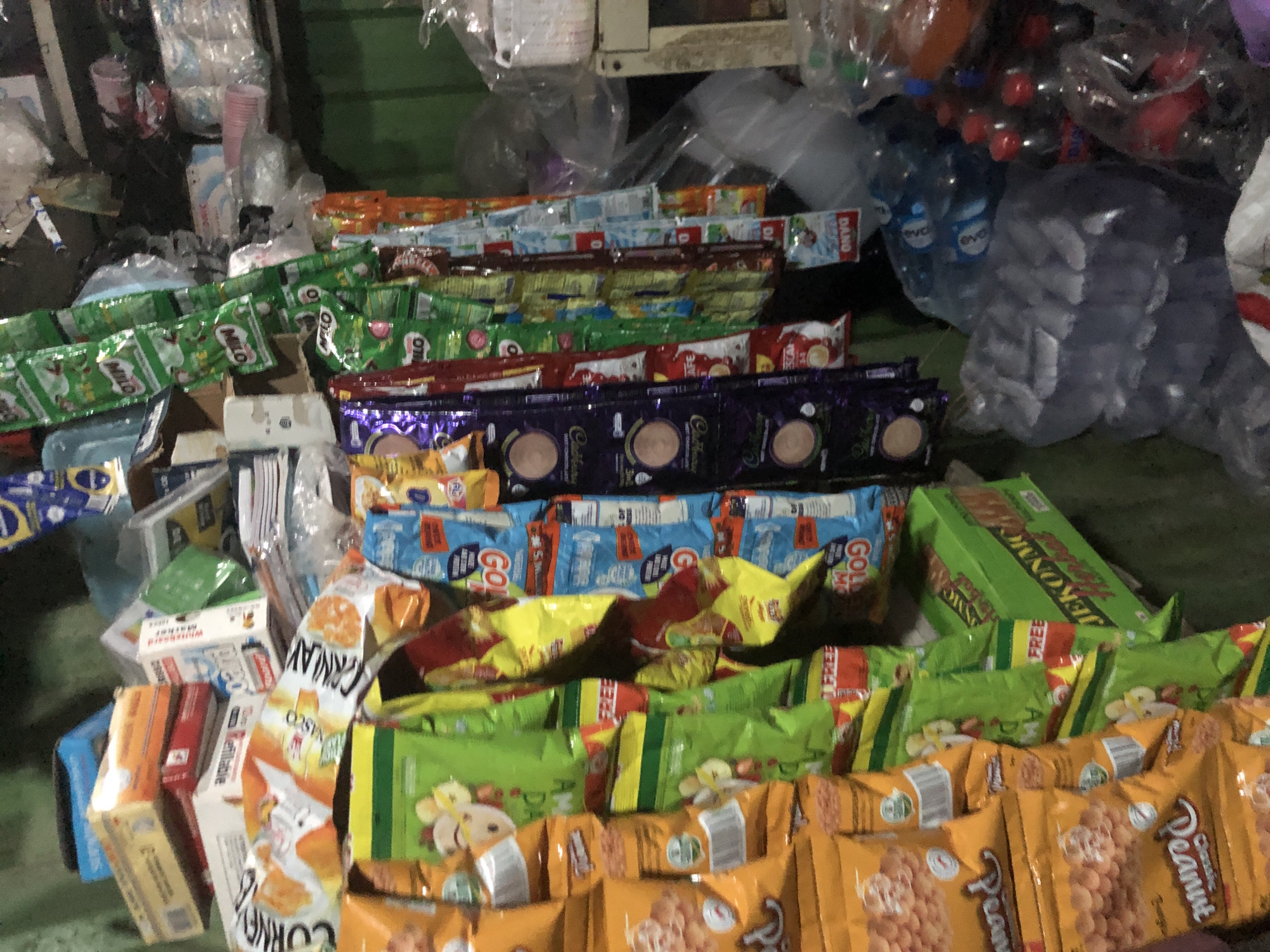The image shows a cluttered shop interior, likely in Ado, Nigeria, filled with various retail goods. Prominently displayed are numerous snack packages, candy bars, and packets of instant beverages, some hanging from racks and others stacked on shelves. There are also bottles of drinks, including water and what appear to be soft drinks, some in clear plastic packaging. Boxes of goods are stacked in the background, and some items are in plastic bags. The overall impression is of a small convenience store or kiosk with a wide array of packaged products for sale. The lighting is somewhat dim, suggesting an indoor, possibly artificial, light source. The environment appears somewhat disorganized, with items piled up in various areas.