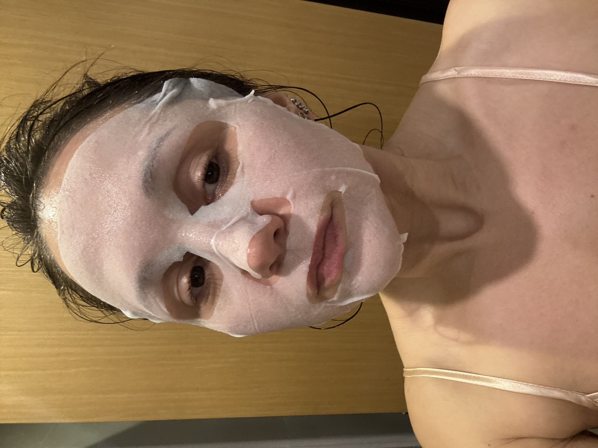 A woman is shown in a close-up shot wearing a facial sheet mask. The mask covers most of her face, with cutouts for her eyes, nose, and mouth. Her hair appears wet and is slicked back. She is wearing a spaghetti-strap top. The background consists of a wooden wall and a dark, possibly metallic surface below. There is no visible indication that the location is Dubai, United Arab Emirates.