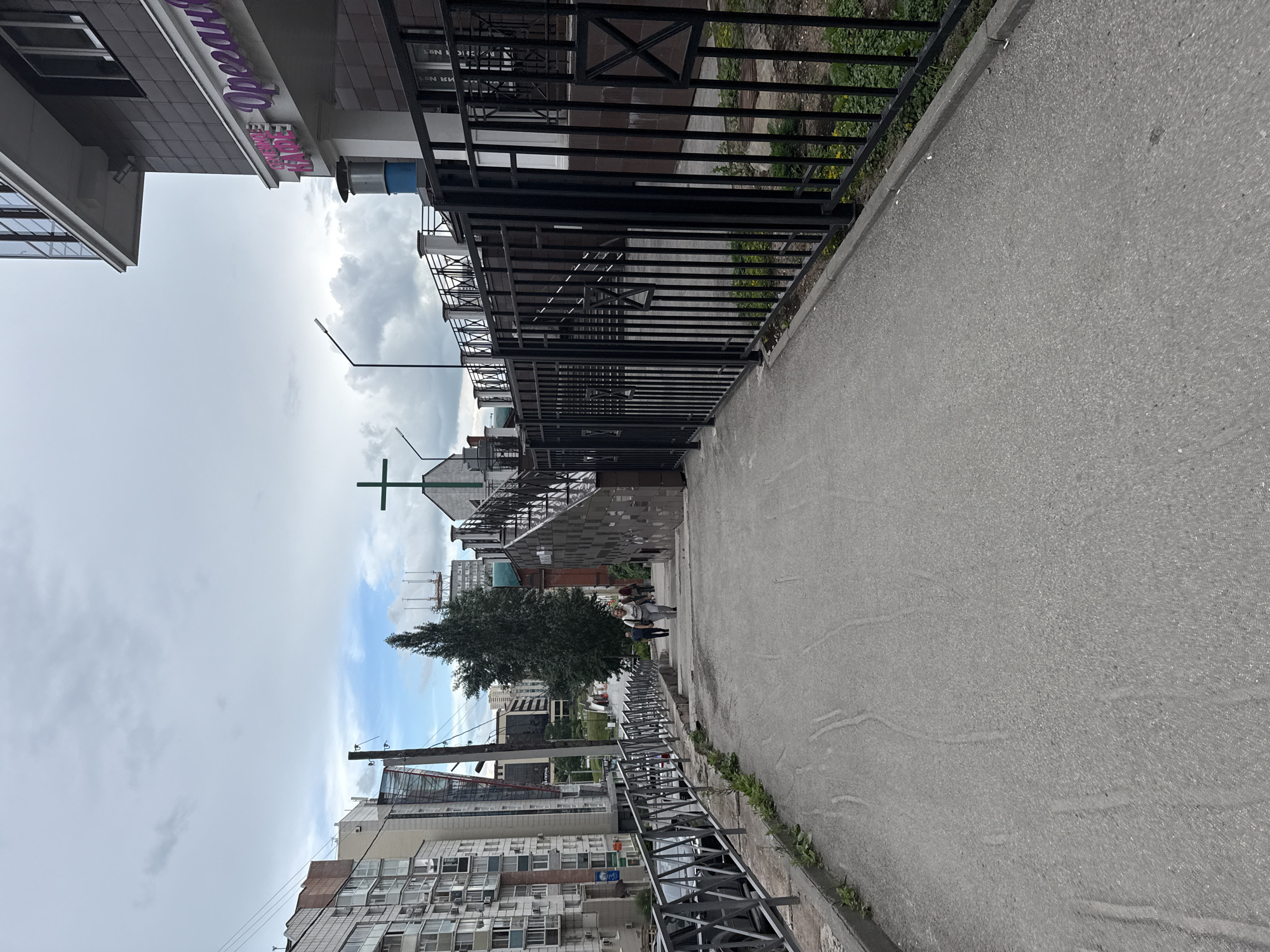 The image shows an outdoor scene on a cloudy day in Perm, Russia. A paved pathway slopes downwards, bordered by a metal fence on the right and a metal railing on the left. Several people are walking down the pathway and up a set of stairs. In the background, there are tall apartment buildings, a large tree, and a green cross structure on top of a building. A sign with Cyrillic lettering is visible on the building to the right. The overall environment appears to be an urban setting with pedestrian walkways and residential structures.