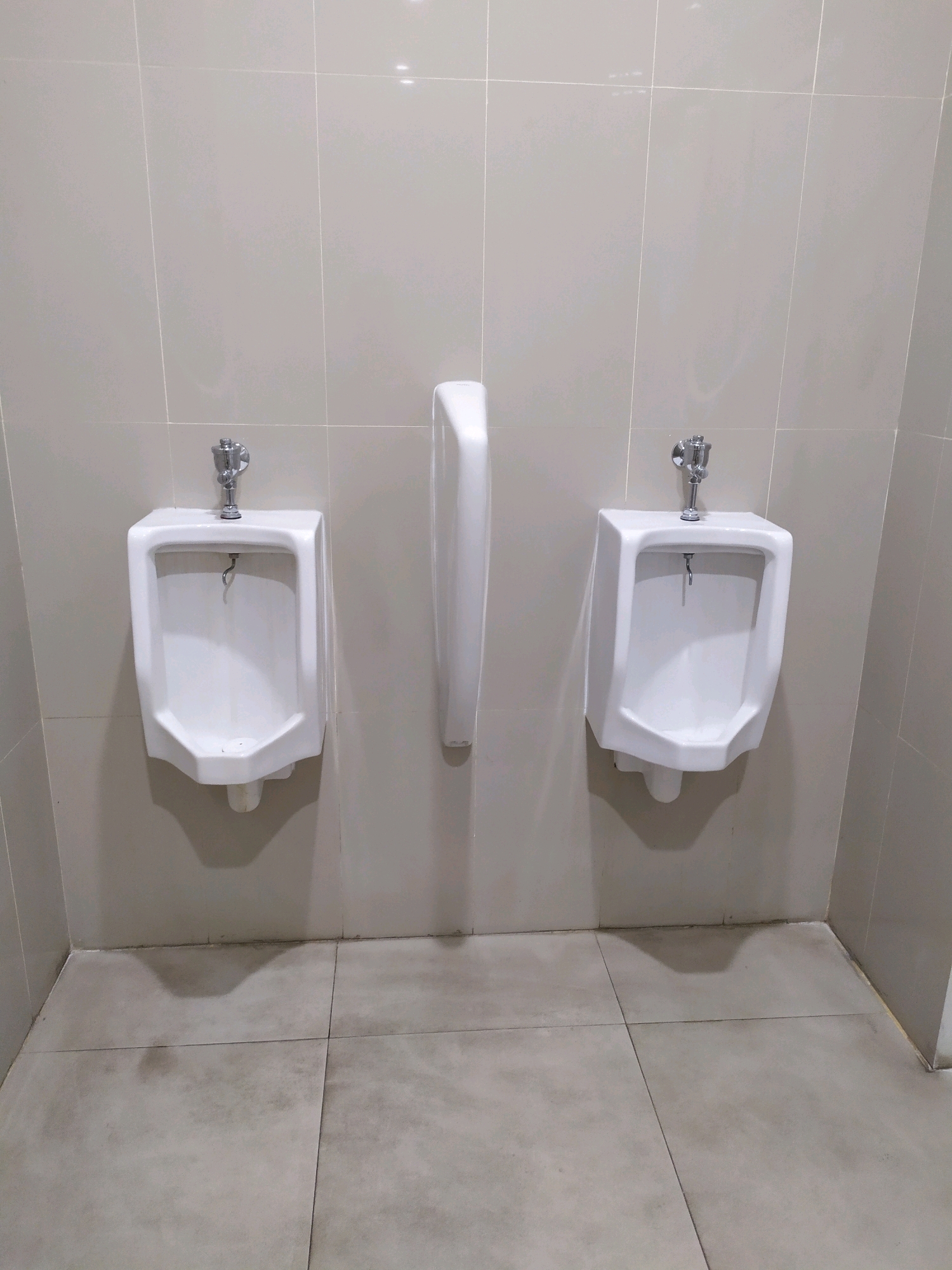The image shows a public restroom in Sleman, Indonesia, featuring two white ceramic urinals mounted on a tiled wall, with a partition in between them. The floor is also tiled, with some damp marks visible. Both urinals have chrome faucets. The overall environment appears to be functional but not recently cleaned.