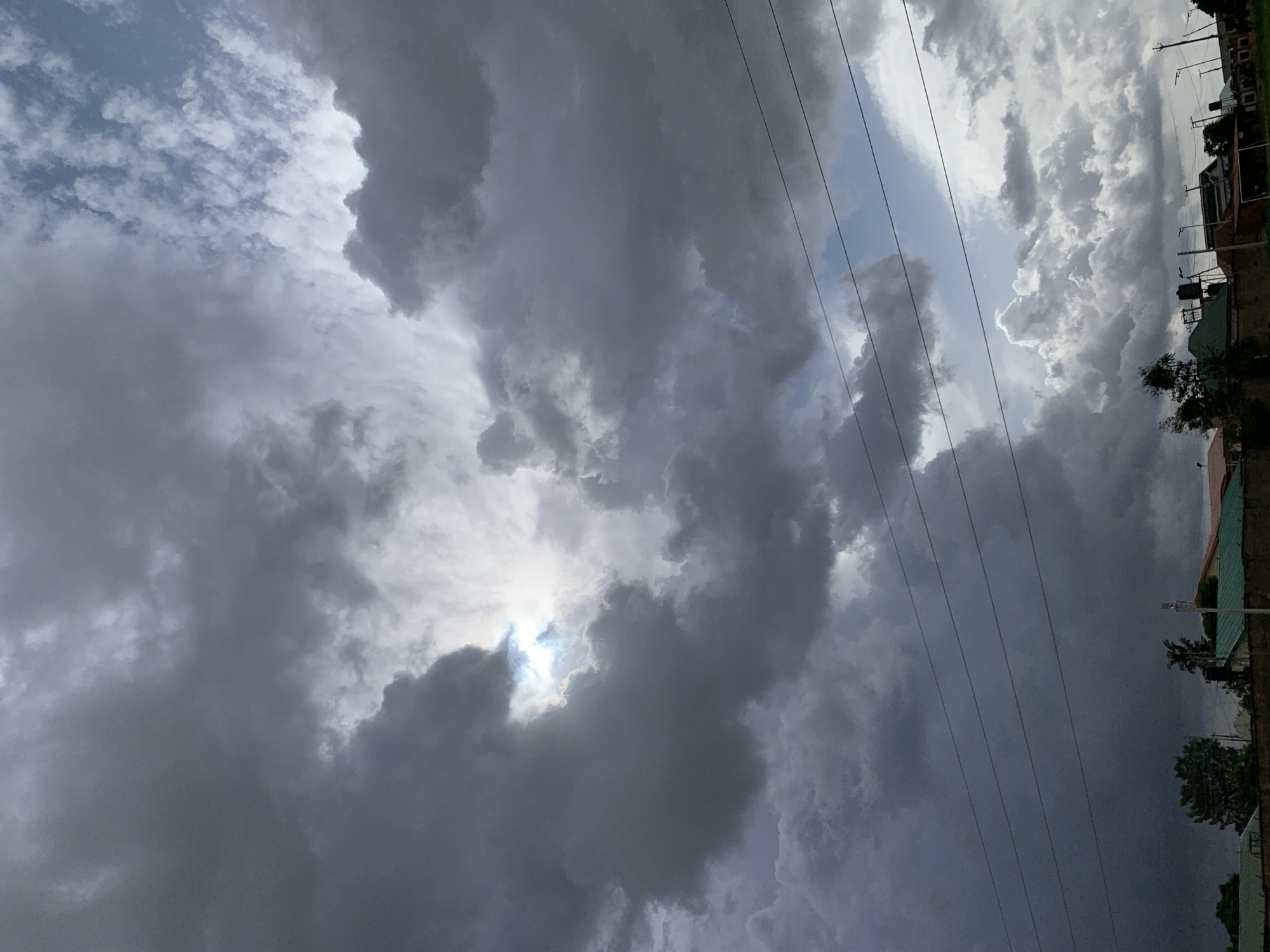 This is a photograph of a cloudy sky over a residential area in Jos, Nigeria. The clouds are a mix of dark storm clouds and lighter, scattered clouds, with a patch of bright sunlight peeking through in the upper center of the image. There are power lines running horizontally across the middle ground of the image, obscuring a view of the buildings below. The buildings appear to be single or two-story homes with pitched roofs. A few trees are visible, as well as some streetlights and a water tank. There are no people visible in the image. The overall scene suggests an impending storm.