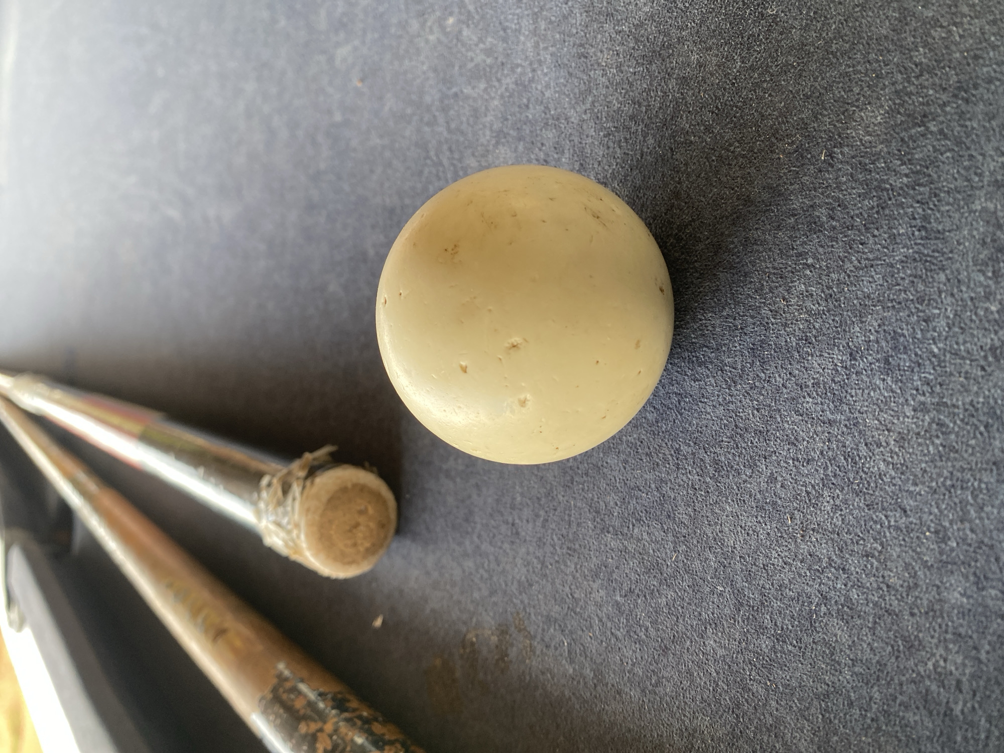 A close-up shot depicts a cue ball resting on a blue pool table surface, with a pool cue resting partially within the frame. The cue ball is off-white and shows signs of wear. The pool cue appears used, with visible texture and color variations. The environment suggests an indoor setting for playing pool. The location in Vom, Nigeria, is not discernible from the image.