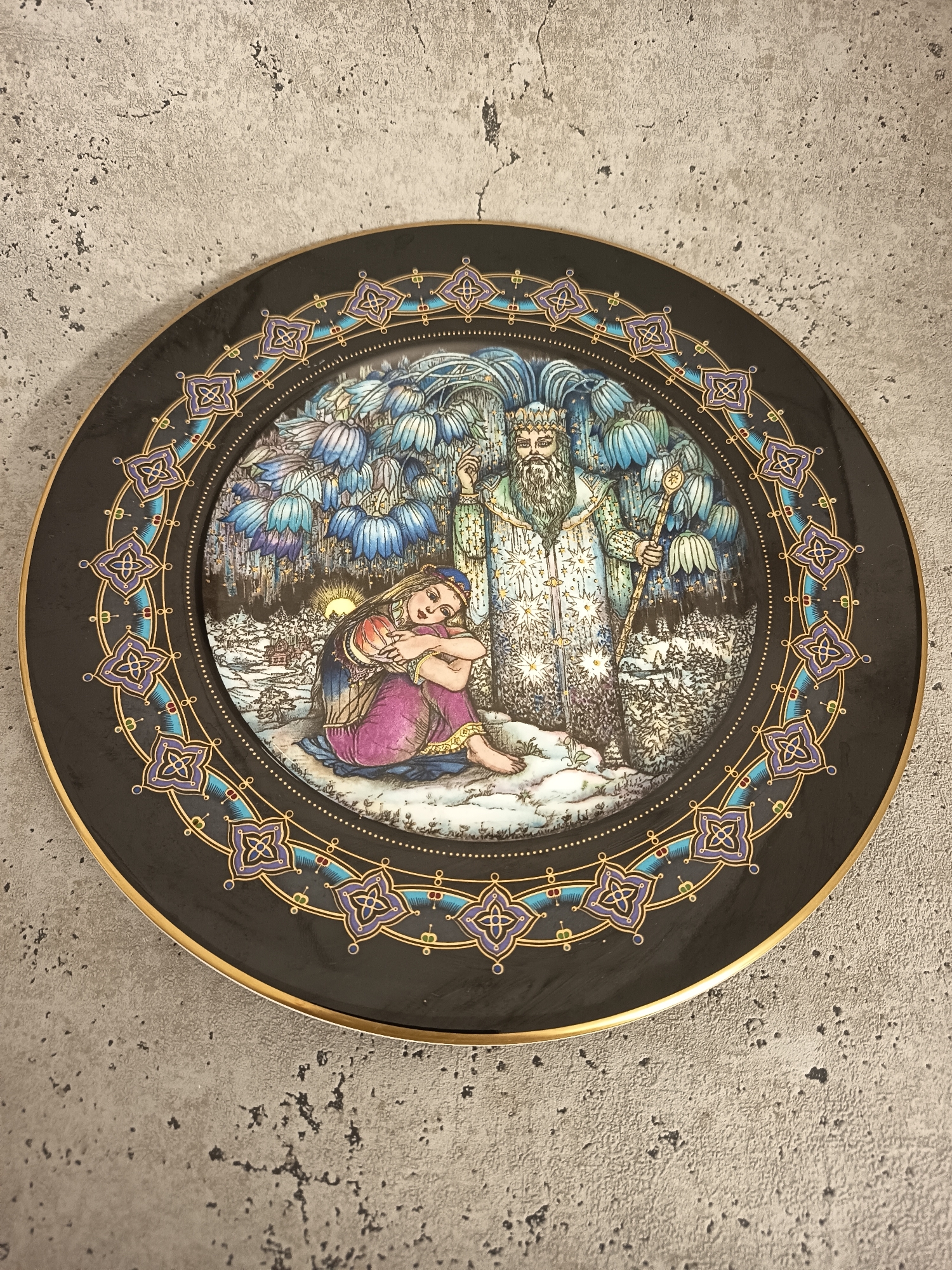 A decorative ceramic plate rests on a grey, textured surface. The plate's center illustration shows a seated woman with long, fair hair, wearing a red top and purple pants, huddled in a snowy landscape with bare feet. Behind her stands a tall, bearded man with a long, star-studded coat and a staff. The background is a forest with blue flowers and a glimpse of the sun. The plate's border features a black background with a gold rim and an inner ring of gold dots, surrounded by a pattern of purple quatrefoils and small colored circles connected by thin gold lines. There is no evidence to suggest the plate is in Wangen bei Olten, Switzerland.