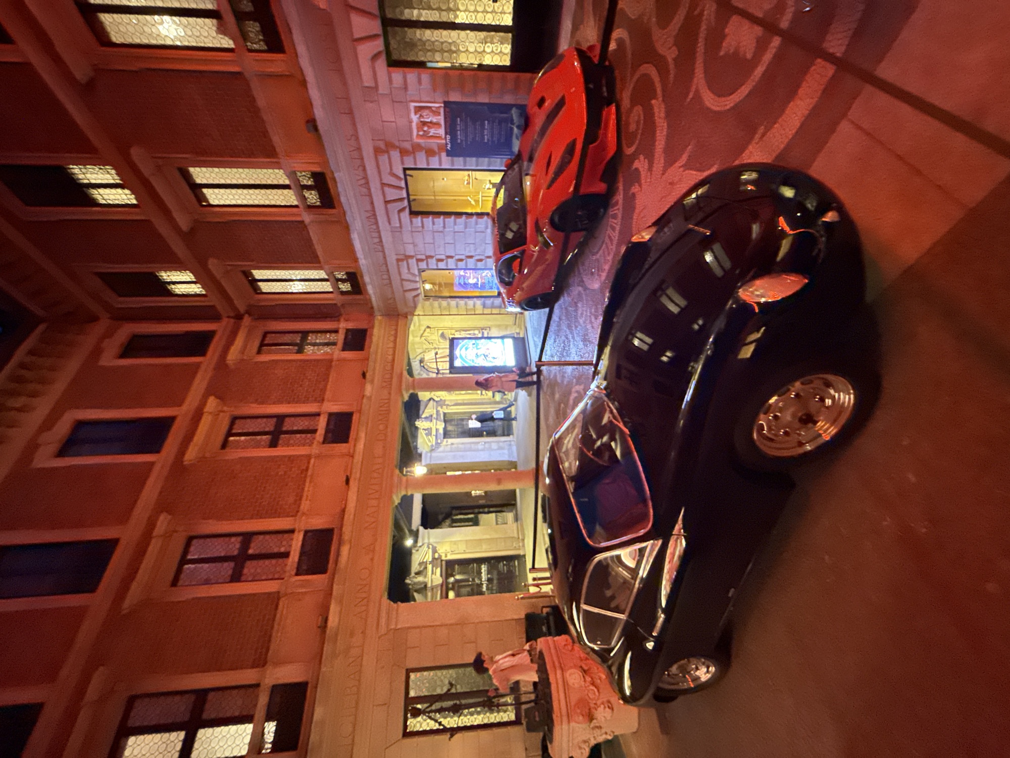 The image captures an evening street scene in an urban European setting, likely Milan, Italy. The time of day is clearly night, with artificial lights illuminating the buildings and street. The overall ambiance is one of sophistication and luxury, characteristic of a high-end district.
The main subjects are two luxury cars parked on the street. In the foreground, positioned diagonally, is a sleek, black, vintage-style convertible car featuring shiny chrome wheels and a polished, reflective body. Behind it, closer to the building facade, a vibrant red, modern sports car with dark wheels is parked.
The scene is set against a backdrop of multi-story buildings with intricate architecture. The facades are adorned with numerous rectangular windows, some showcasing ornate grilles or patterned glass, and several are brightly lit from within. The ground features a decorative patterned paving, possibly cobblestones or ornate tiles, visible around the cars.
Several people are visible in the background, though they are indistinct due to distance and lighting. One figure in a light-colored coat is partially obscured by the black car on the left, near a stone balustrade. Other figures can be seen further down a lit archway or passage between buildings.
Notable details include an elaborate Latin inscription carved into the stone facade of the building on the left, which reads in parts: "...FAVSTVS... PATRVM... ANNO A. NATIVITATE DOMINI MDCCCLXXI" (In the year of the Lord's nativity 1871), providing a historical context to the architecture. Near the red car, a sign or poster is visible with the partial text "AUTO". The warm, reddish-orange glow of the lighting creates a dramatic and inviting atmosphere. There is no specific activity or event taking place, rather it depicts a bustling street during the evening.