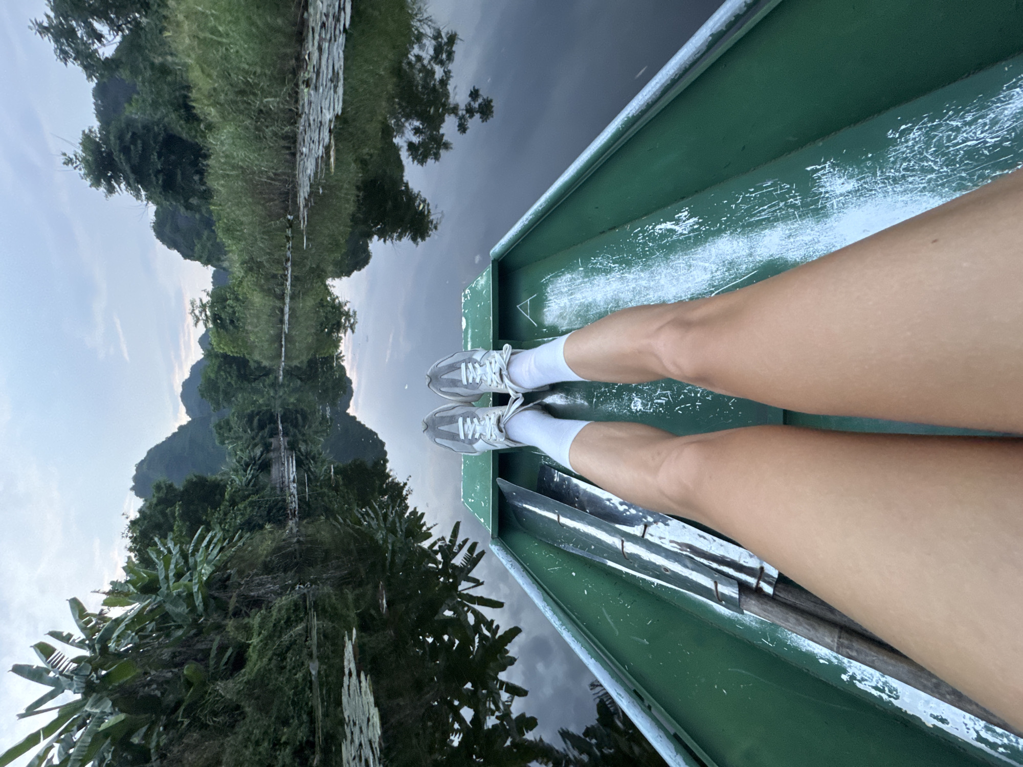The image shows a first-person perspective of someone sitting in a green boat on a body of water, likely a river or canal, in Nam Hoa Lư Ward, Vietnam. The water is dark and reflects the surrounding lush vegetation and the sky. In the foreground, the person's legs, wearing white socks and grey sneakers, are visible, stretched out in the boat. A paddle rests alongside the person's left leg. The boat itself appears to be made of metal and shows signs of wear, with white paint chipping away.
The environment is characterized by dense tropical foliage lining the water's edge, including large-leafed plants that appear to be banana trees, as well as tall reeds and other greenery. In the background, karst mountains rise above the trees, their slopes covered in vegetation. The sky is overcast, with hints of twilight. The overall impression is one of a serene and natural landscape, possibly a tourist excursion through a scenic waterway.