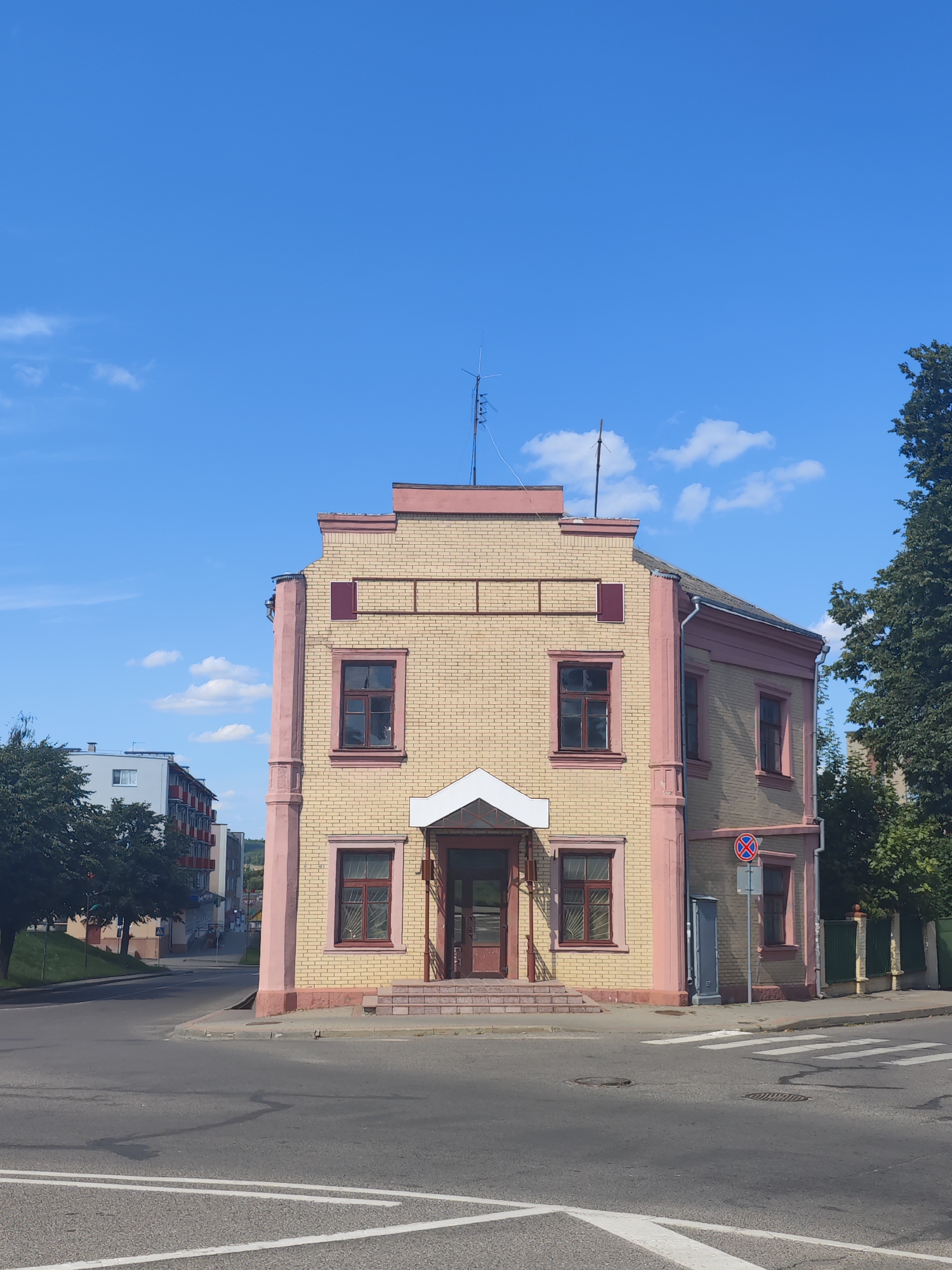 The image shows a two-story yellow brick building with pink trim, located at a street intersection in Slonim, Belarus. The building has multiple windows with dark frames and a small porch over the entrance. A pedestrian crossing is visible in the foreground. To the left of the building, there is a modern multi-story residential building and some trees. To the right, there is a tree and a green fence. The sky is blue with scattered white clouds. The scene appears to be a sunny day.