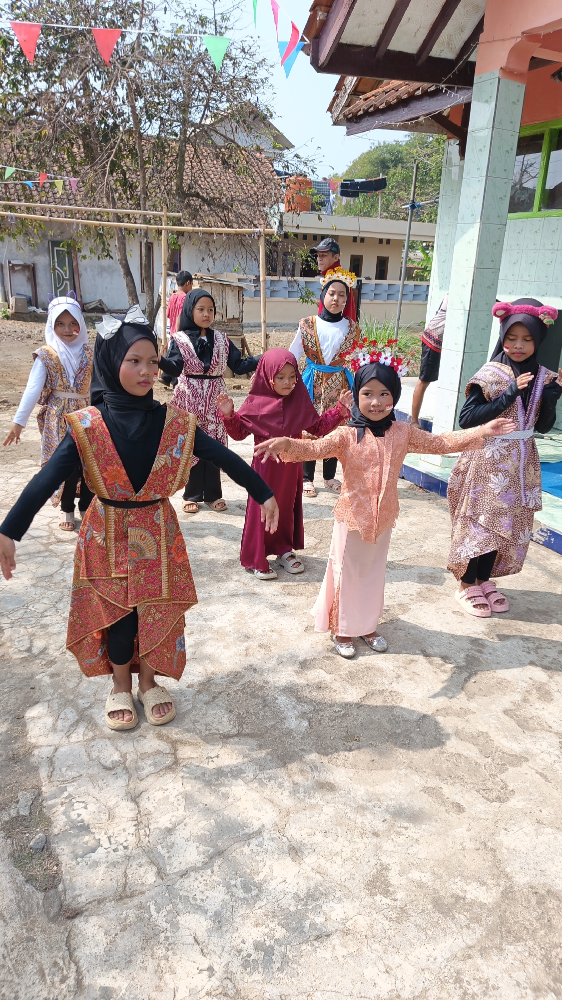 In a village setting, likely in Linggasirna, Indonesia, a group of young girls, possibly participating in a cultural event or performance, are captured in a candid outdoor scene. The ground is a cracked concrete or asphalt surface, suggesting an unpaved or rustic courtyard. In the background, a building with a distinct tiled roof and greenish-blue tiled columns stands, hinting at a local community structure. Lush green trees and other vegetation add to the rural ambiance. Decorative flags, red and green, strung on a bamboo pole, suggest a festive occasion. The girls are dressed in colorful, traditional outfits. Several wear headscarves, varying in color from black to white and maroon. Their clothing features intricate patterns and designs, indicating a cultural or traditional attire. Some girls have face paint, with red markings on their cheeks. They appear to be in motion, with their arms outstretched, suggesting they are either dancing or posing for a photograph. One girl in the foreground wears a striking red and gold patterned tunic over black leggings, accessorized with woven sandals. Another girl, to her right, is dressed in a peach-colored traditional top and skirt, adorned with a floral headdress. The overall scene conveys a sense of community celebration and cultural pride in a traditional Indonesian village environment.